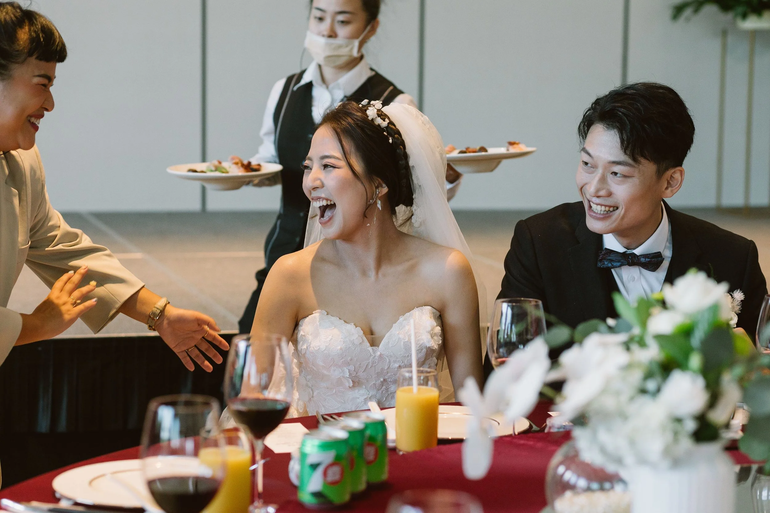 A bride and groom, both smiling, seated at a table during a celebration, with a woman in a beige outfit, another woman in a white shirt and black vest, and a woman in a wedding dress, all smiling and talking. The table has wine glasses, drinks, and a