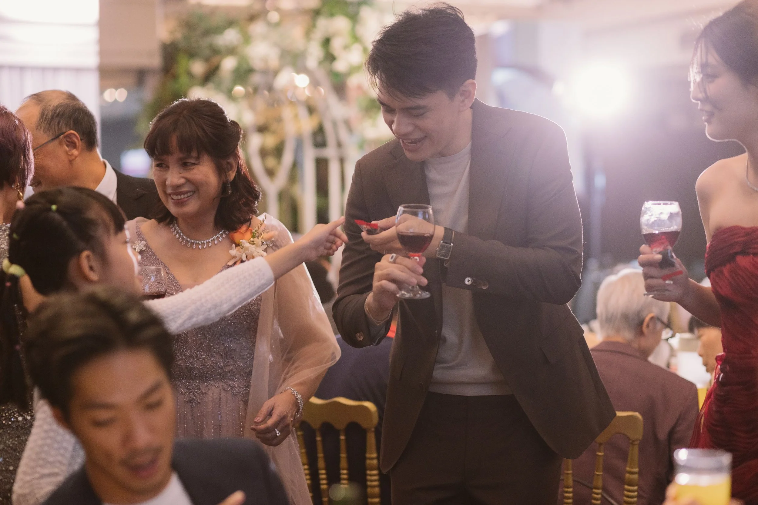 People at a social gathering, with one man smiling and holding a glass of red wine, engaging in conversation with a woman. The scene appears lively with warm lighting.