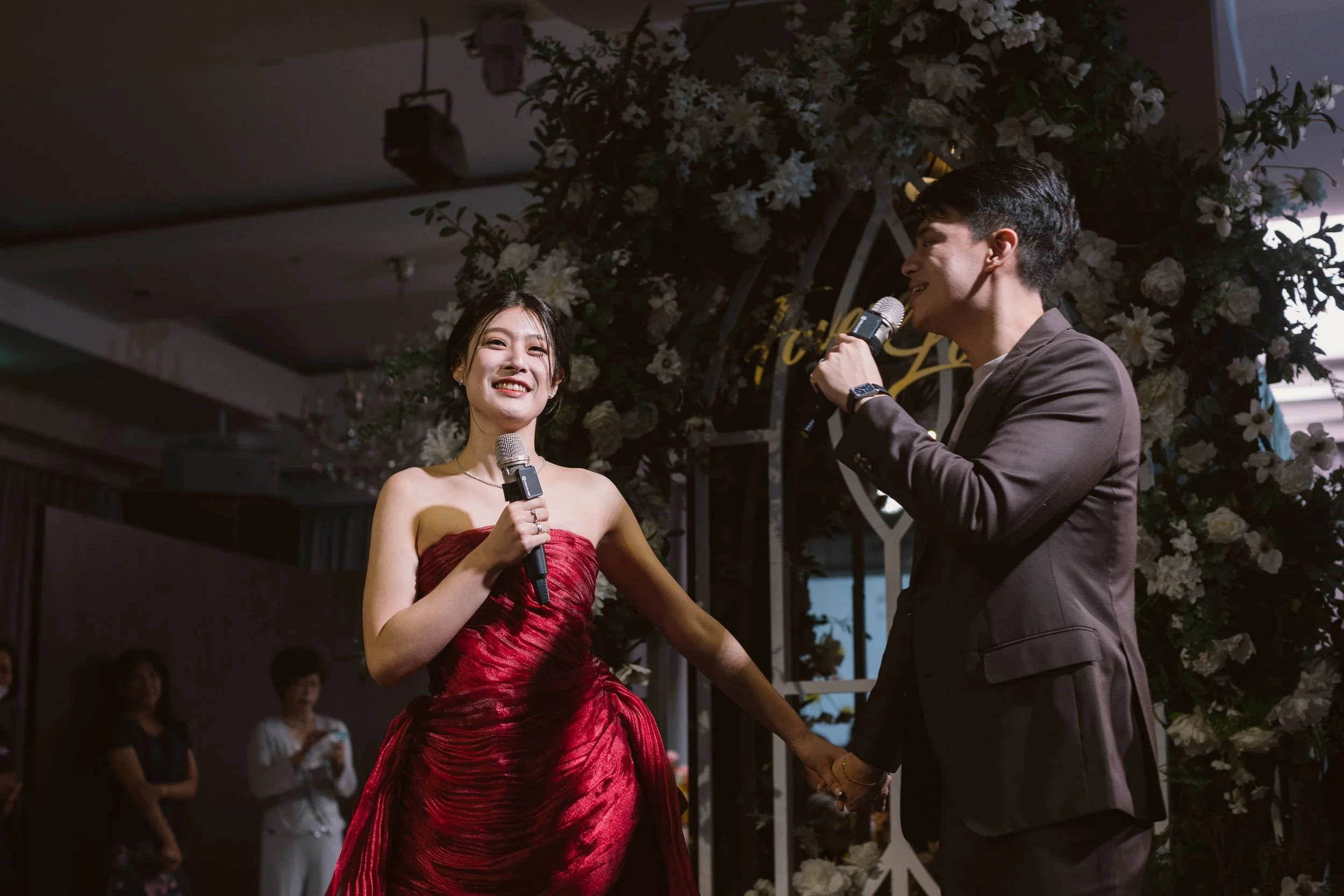 A woman in a red strapless gown smiling, holding a microphone, standing next to a man in a dark suit with a microphone, at a formal event with floral decorations and audience members in the background.
