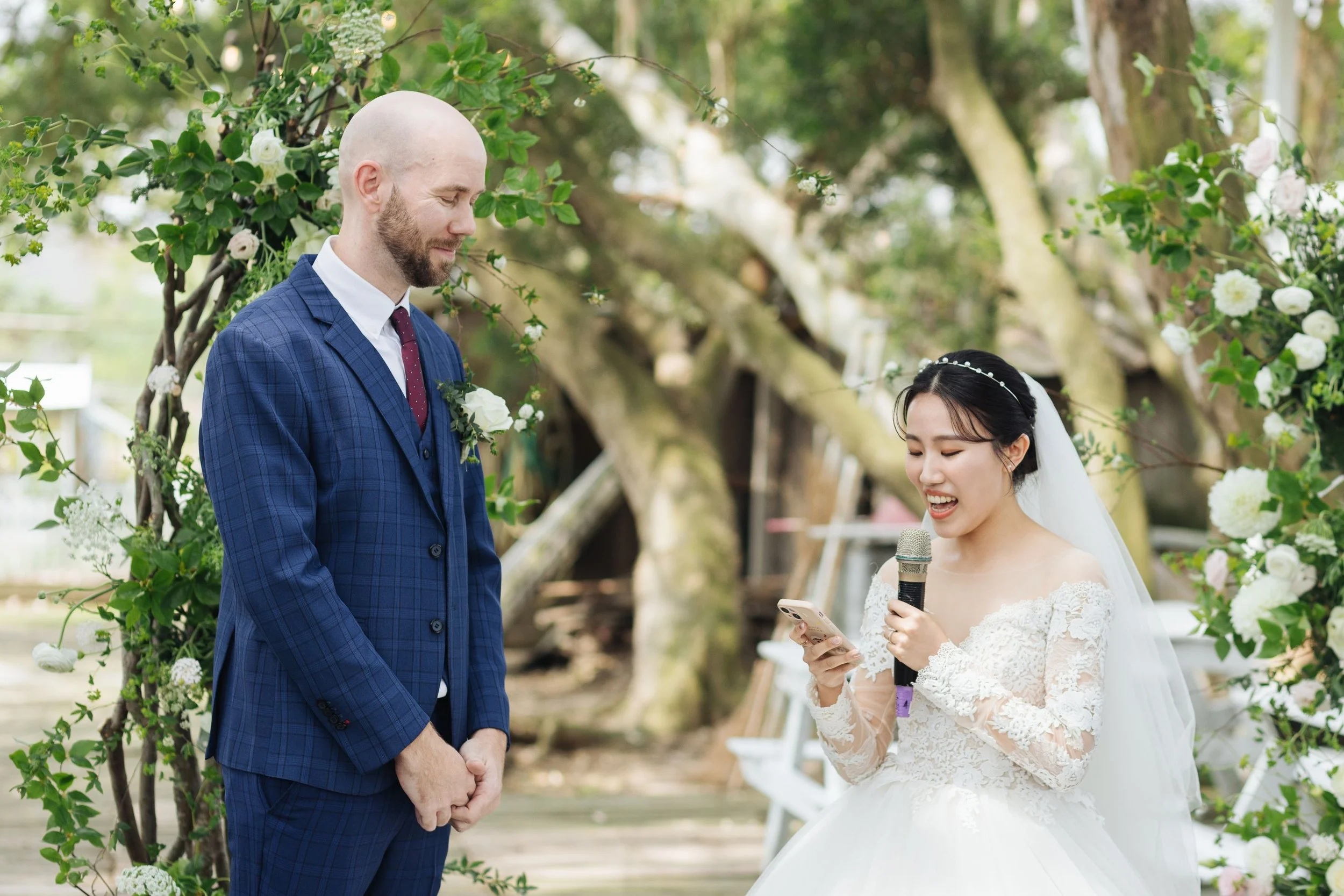 A man and woman at a wedding ceremony outdoors, with the woman reading from a phone and holding a microphone, surrounded by greenery and flowers.