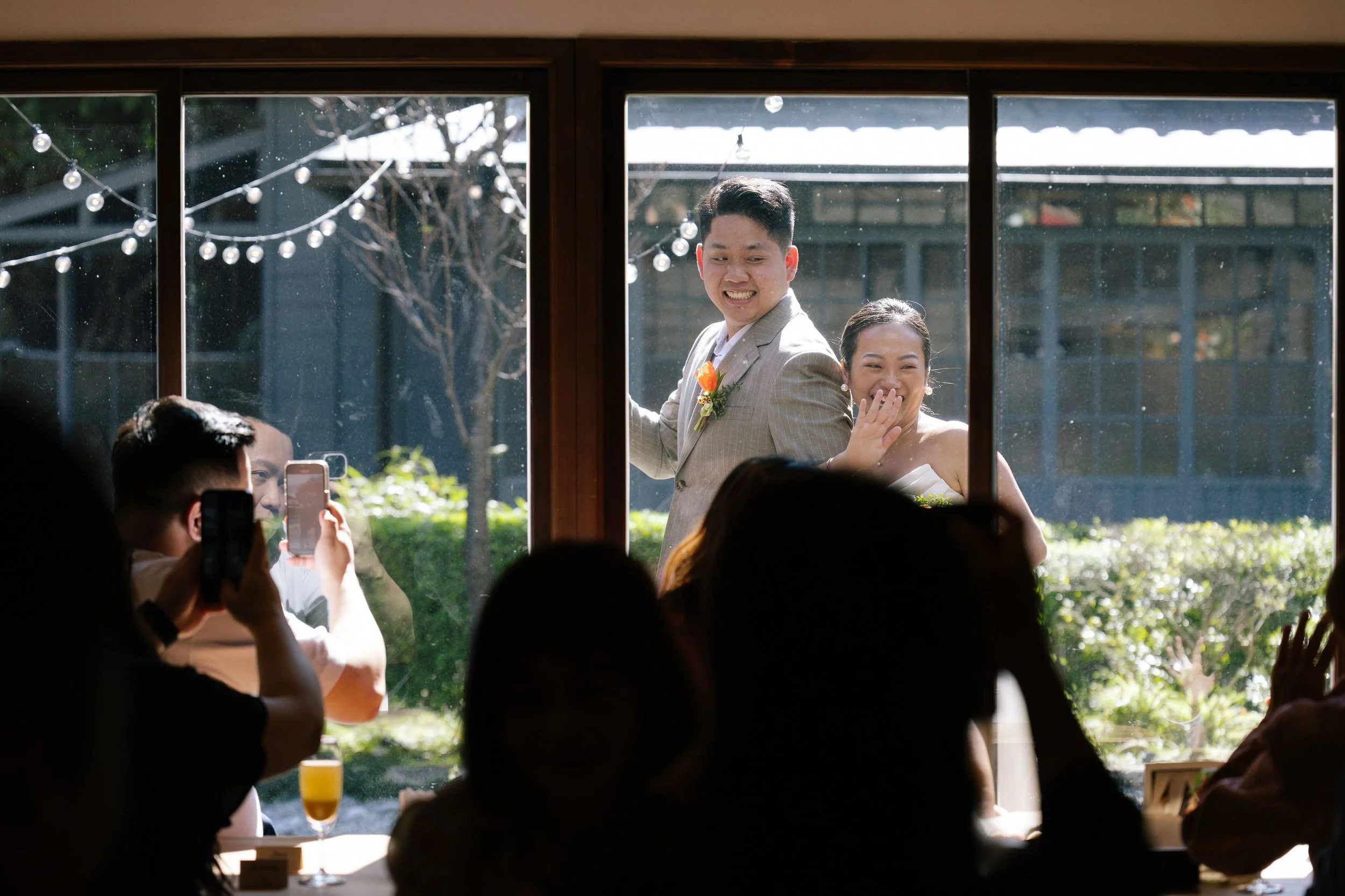 View through a window showing a smiling couple, a man in a suit with a boutonnière and a woman in white, at a celebration, with guests inside taking photos, and string lights outside.