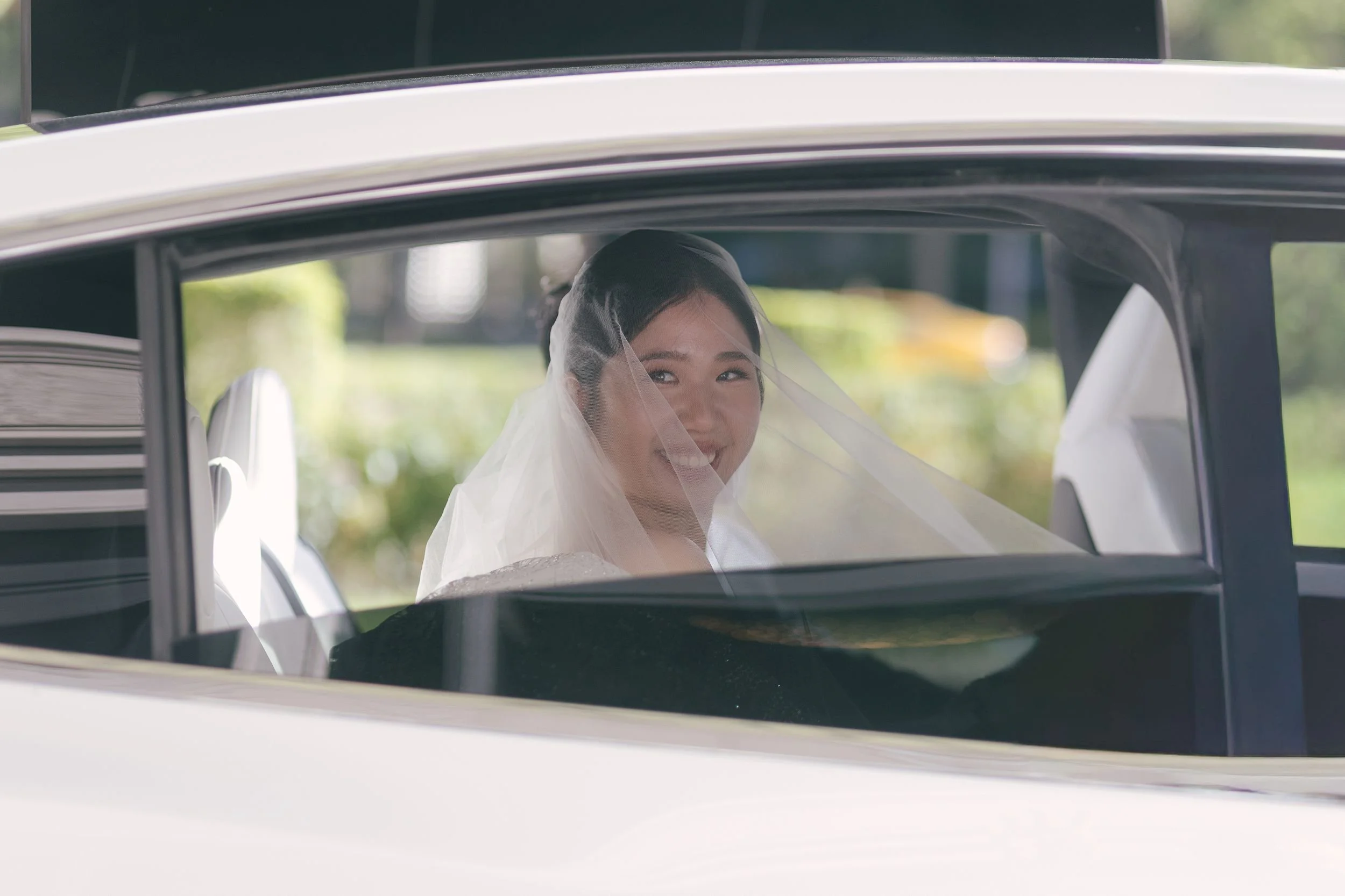A bride with a veil smiling and looking out the window of a car.