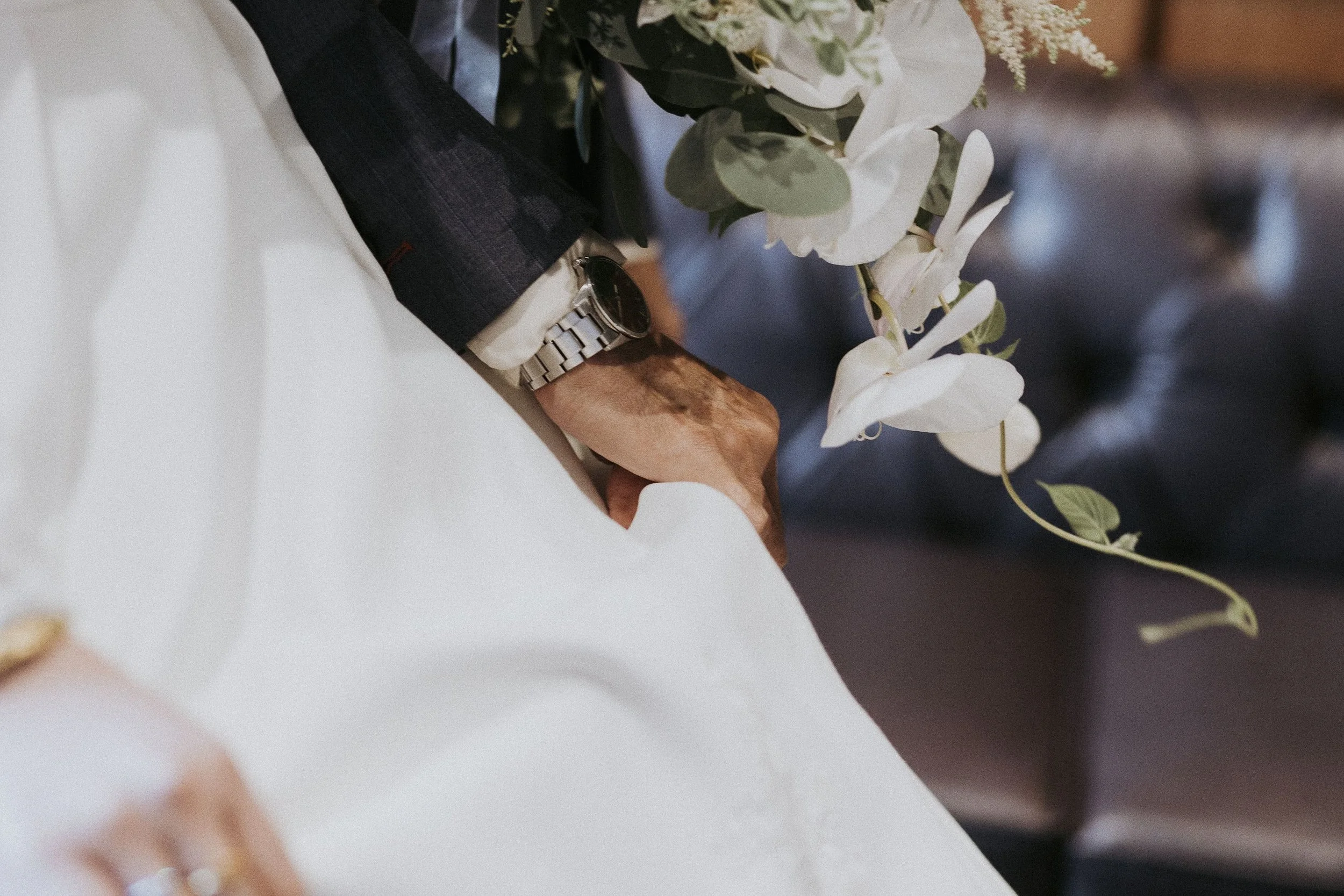 Close-up of a person's arm wearing a wristwatch and a dark-colored suit sleeve, resting on a white surface beside a bouquet of white and green flowers.