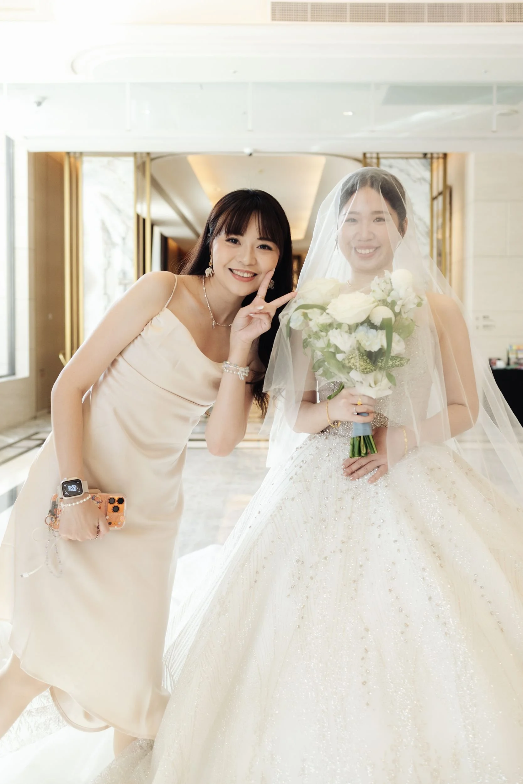 A woman in a champagne-colored dress smiling and making a peace sign next to a bride in a white wedding gown holding a bouquet of white flowers, both posing indoors in a well-lit, elegant setting.