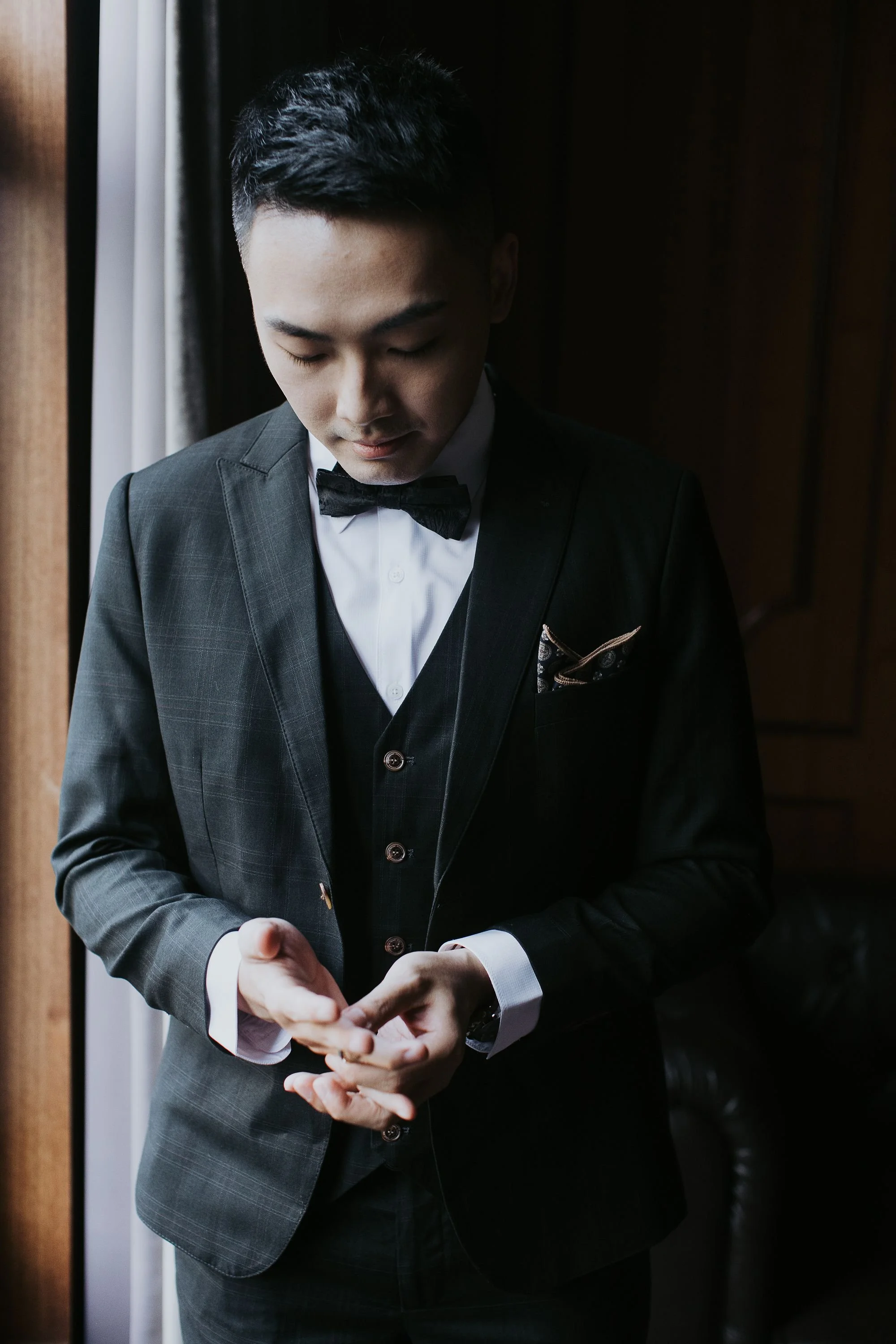 A man in a black tuxedo with a bowtie, holding a smartphone in his hands, standing near a window with curtains.