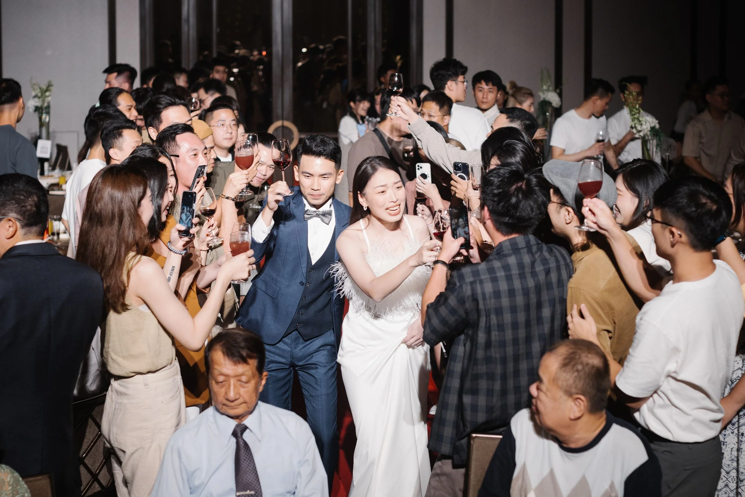 A wedding reception with many people holding glasses of wine and taking photos, with a couple, possibly the bride and groom, in the center celebrating.