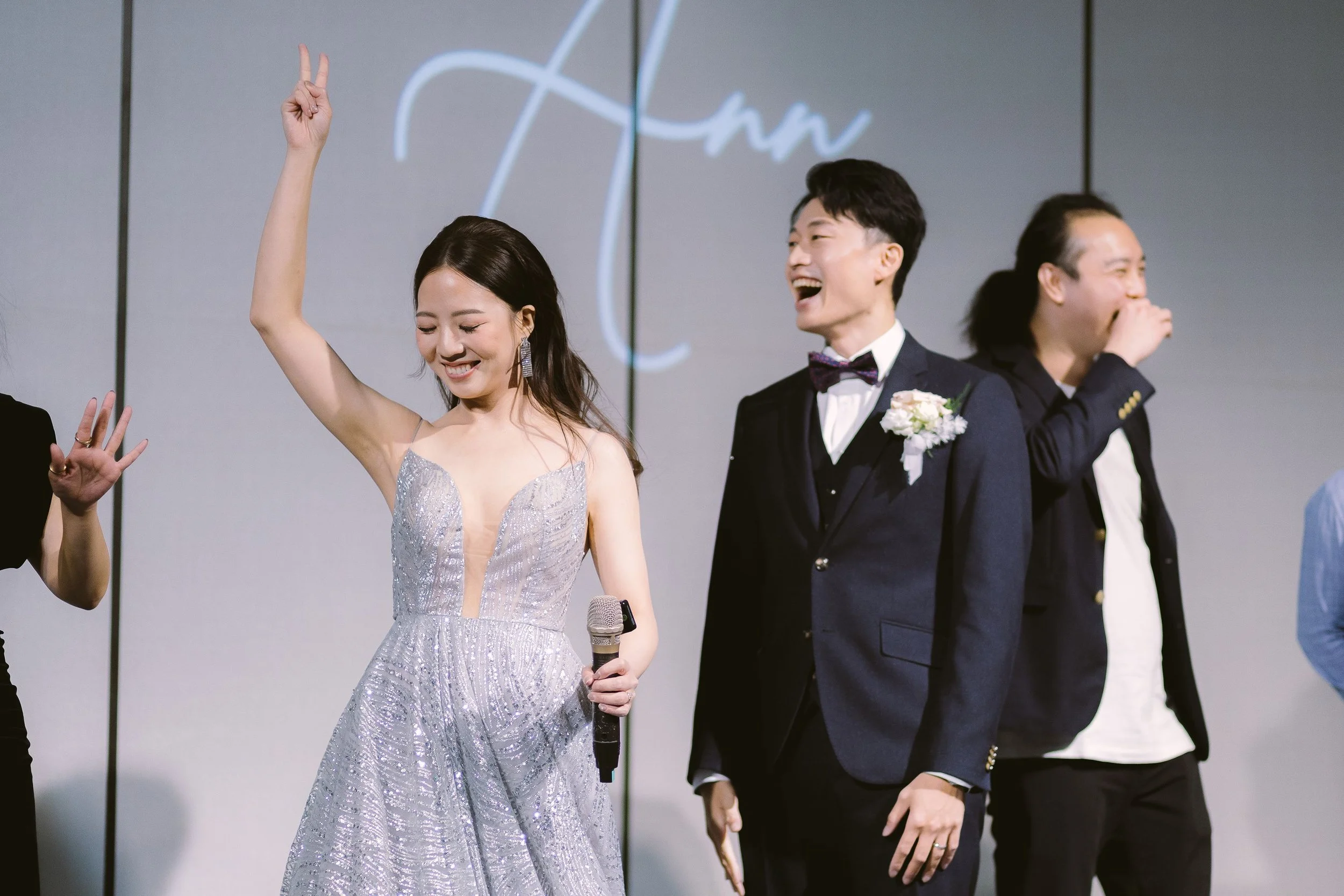 A woman in a silver dress holding a microphone stands on stage, smiling and making a peace sign with her hand. Two men in suits, one with a bow tie and corsage, are laughing and smiling nearby. The background features a large screen with stylized wri