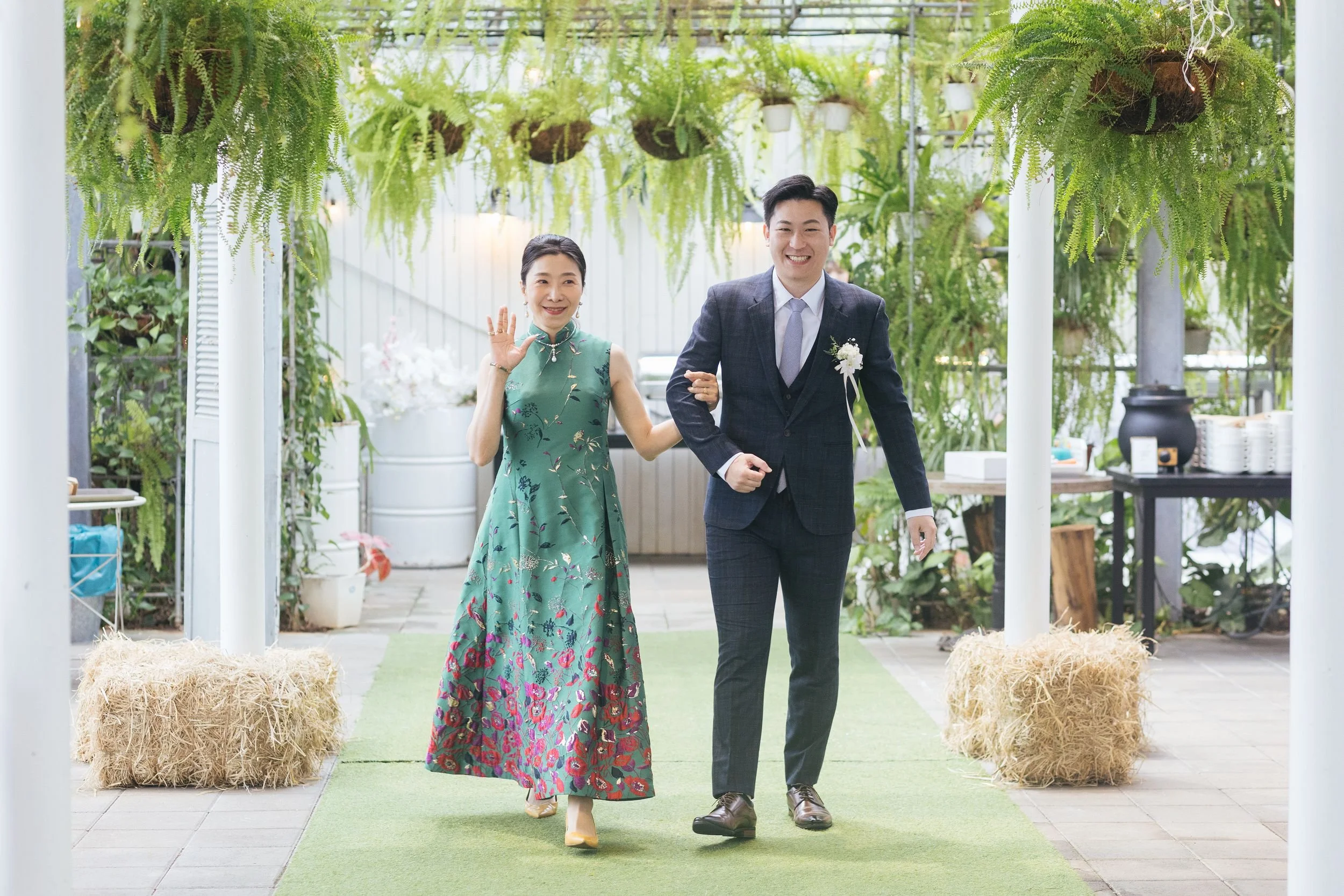 A groom in a dark plaid suit and a bride in a colorful floral dress walking together in a greenhouse decorated with hanging ferns and potted plants, smiling and waving.