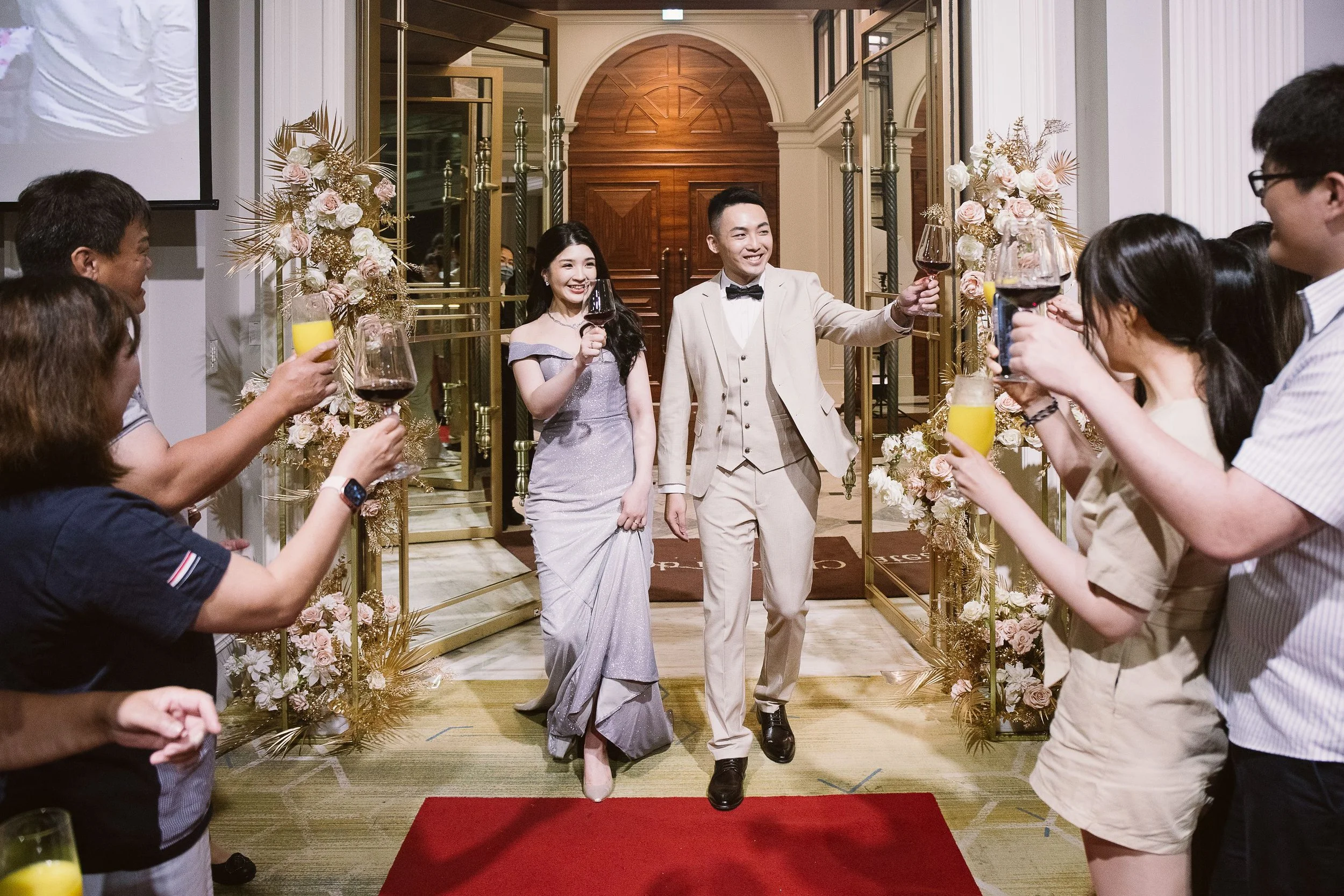 A couple in formal attire standing on a red carpet, holding glasses of red wine, surrounded by guests at a wedding celebration. The woman is wearing a silver evening gown and the man is in a beige suit with a black bow tie, smiling and toasting. The 