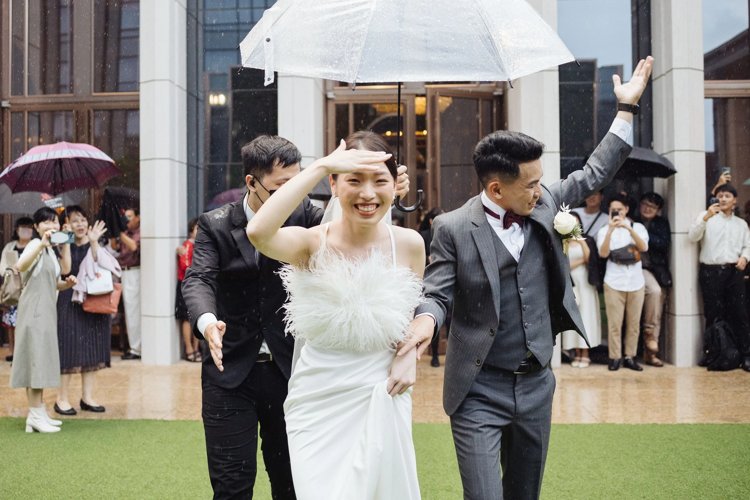 A bride and groom celebrating outdoors in the rain with a crowd of friends taking photos and cheering in the background. The bride is smiling and holding a feathered clutch, while the groom raises his hand.