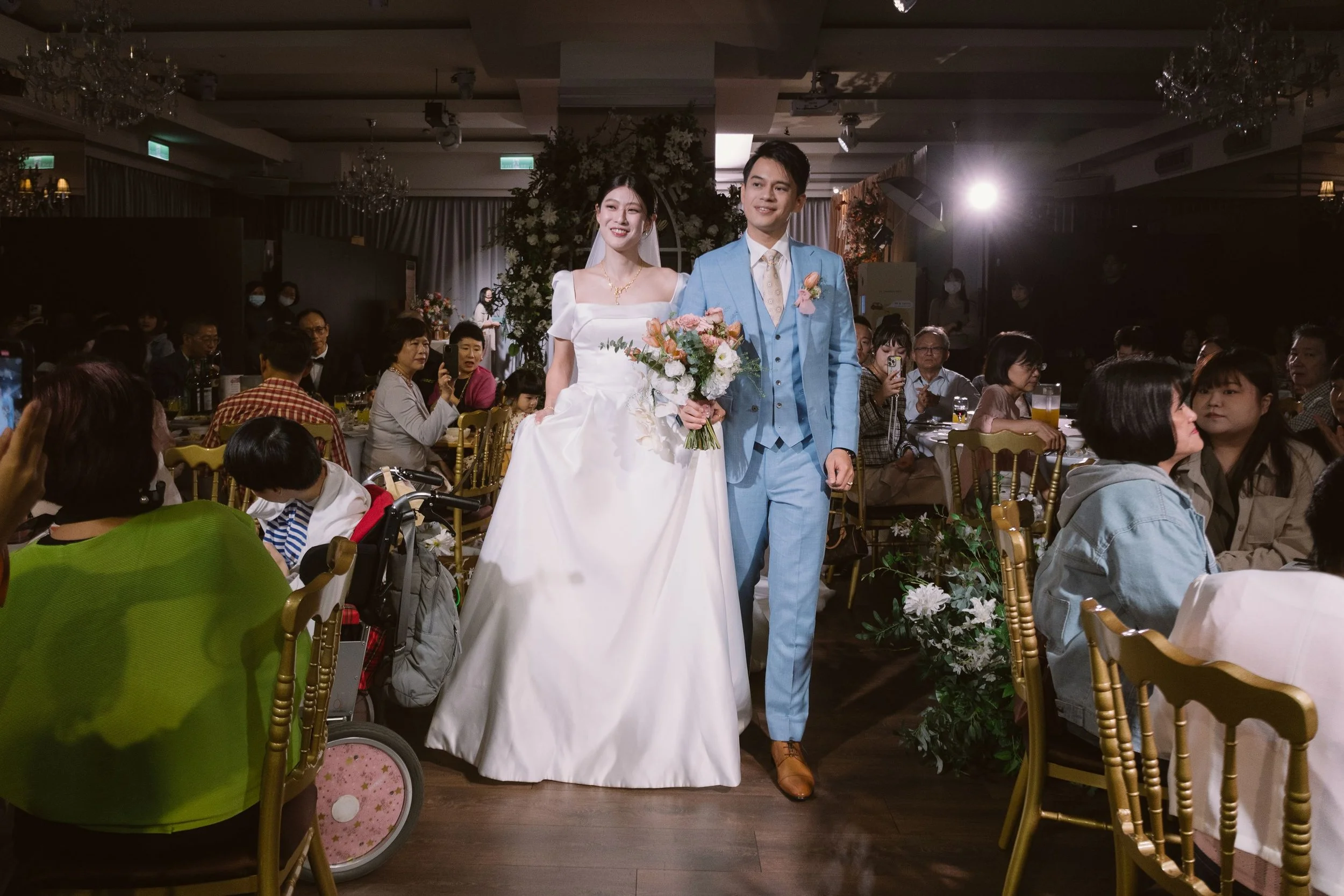 A bride and groom walking down the aisle at their wedding reception, surrounded by seated guests in an indoor venue with chandeliers and floral decorations.