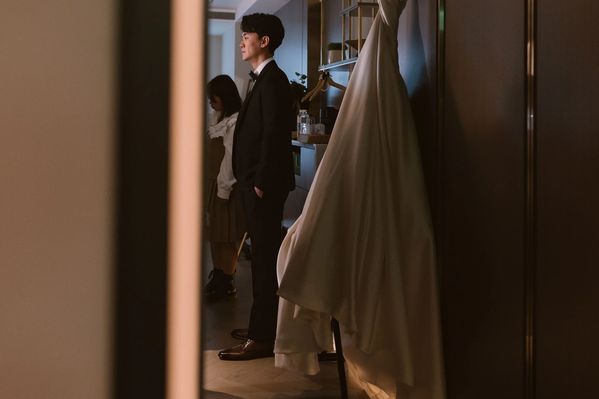 A man in a tuxedo and a woman in a dress standing in a room, seen through a narrow opening or gap, with a draped fabric or curtain in the foreground on the right side.