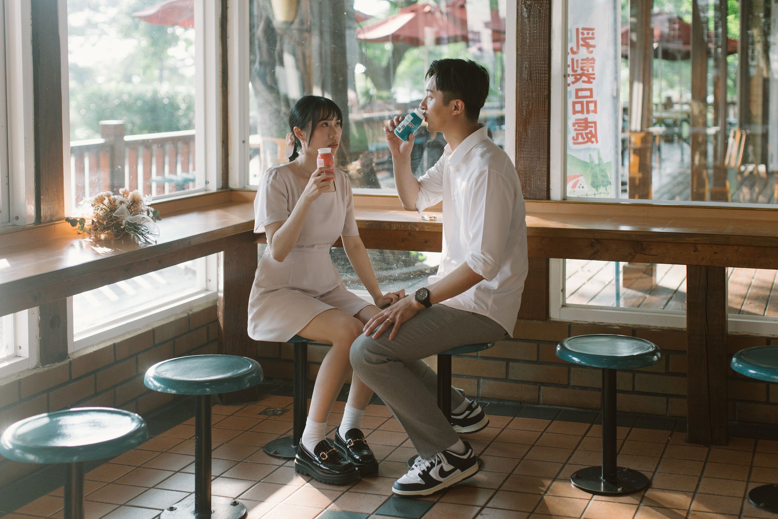 A young man and a young woman sit at a counter in a restaurant, drinking from bottles. The woman looks at the man who is drinking. There is a bouquet of flowers on the window sill beside them. Large windows with a view of trees and outdoor seating ar