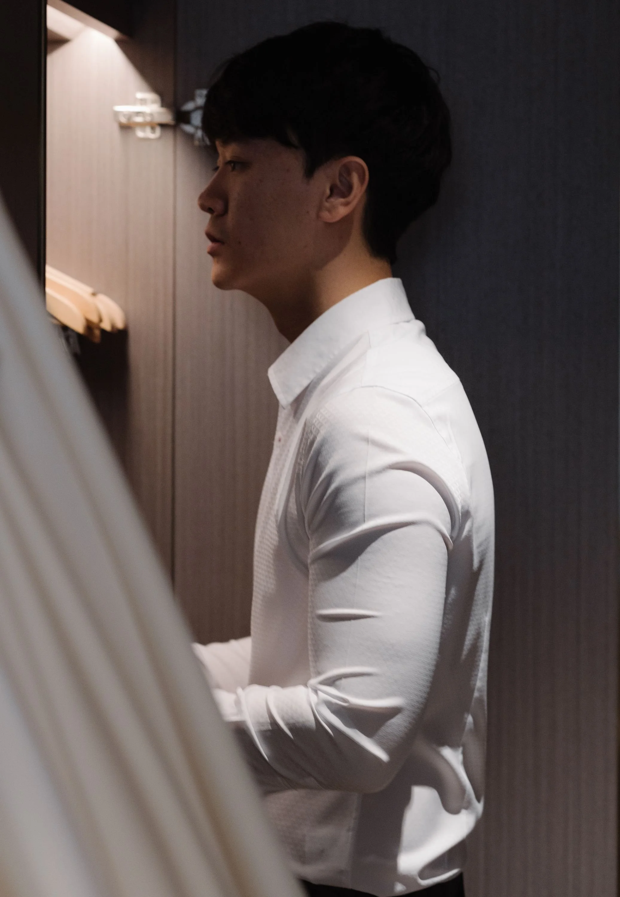 A young man with dark hair in a white shirt is sitting with his back straight, looking forward. He is against a dark wall, near a wooden wardrobe with hangers, in a dimly lit room.