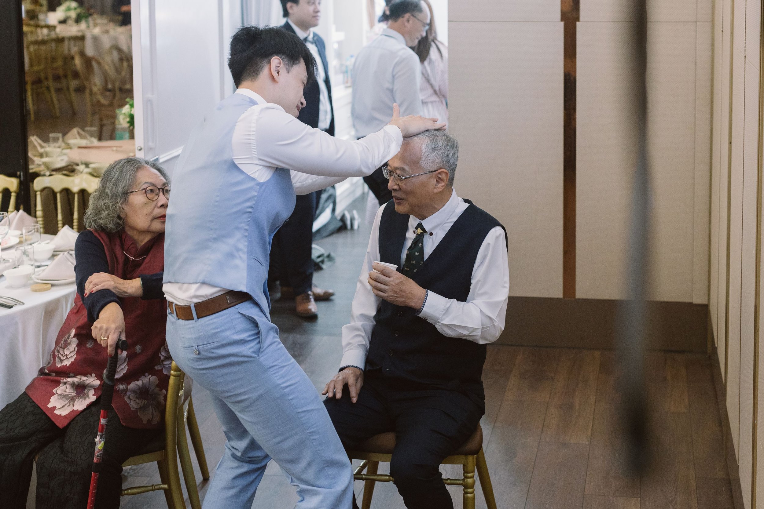 A group of elderly and middle-aged people at a social gathering or celebration, with one man receiving a physical blessing or comforting gesture from a younger man. The scene includes a dining area with tables set with dishes and glasses, and people 