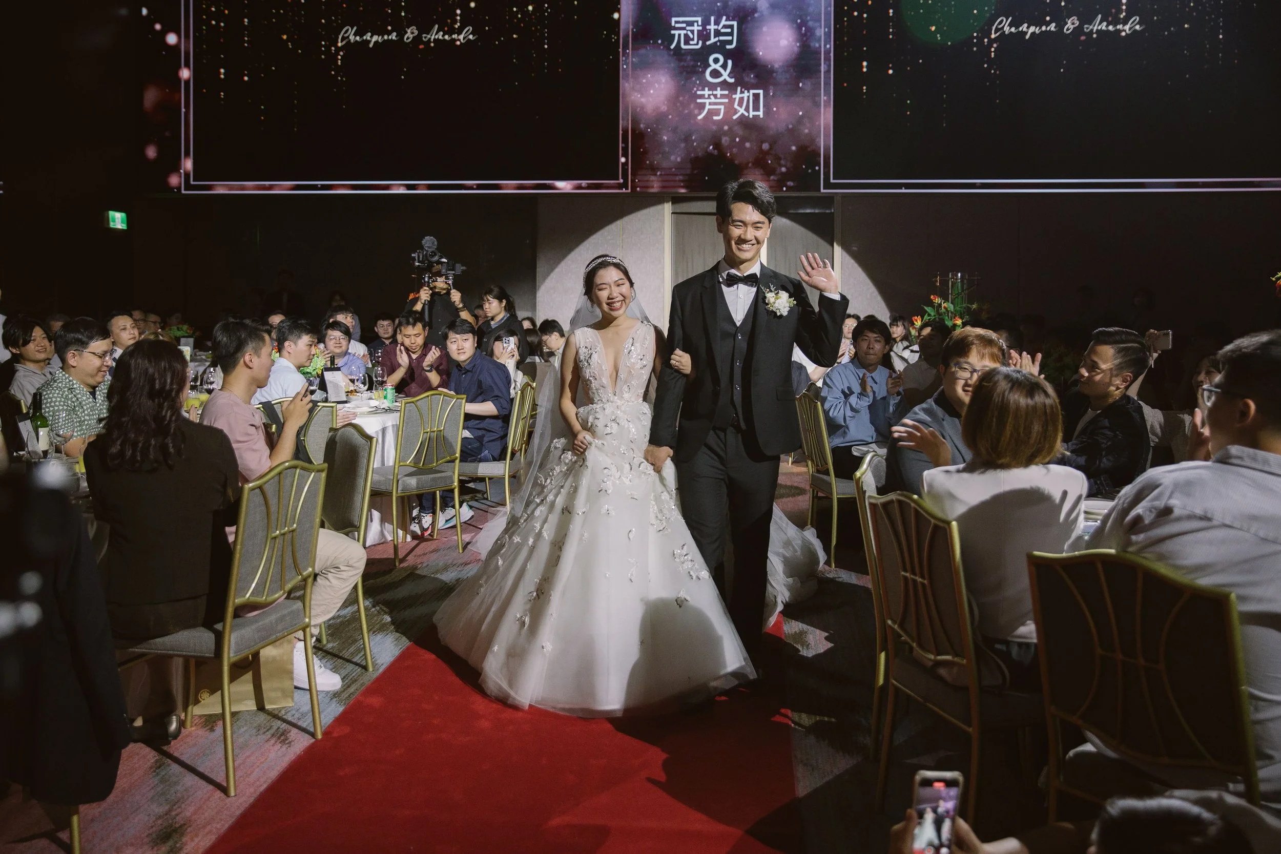 A bride and groom walking down a red carpet at their wedding reception, surrounded by seated guests. The bride wears a white wedding gown with floral embellishments, and the groom wears a black tuxedo with a bow tie. Guests are clapping and smiling a