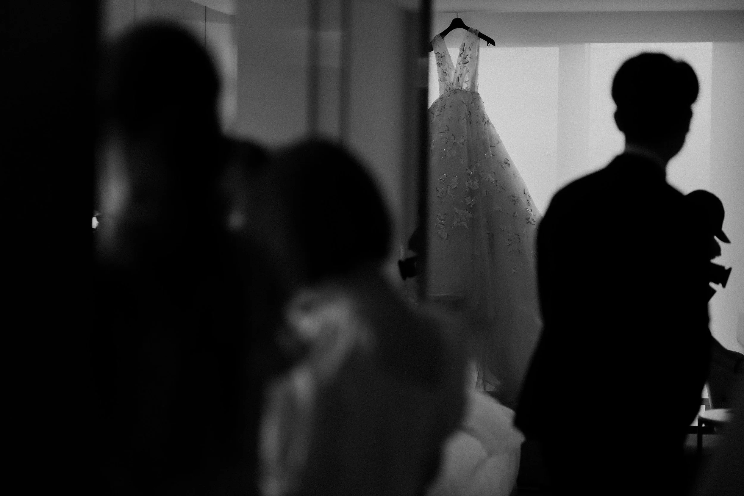 Silhouetted figures of a man in a suit and a woman in a dress, with a wedding gown hanging on a hanger in the background.