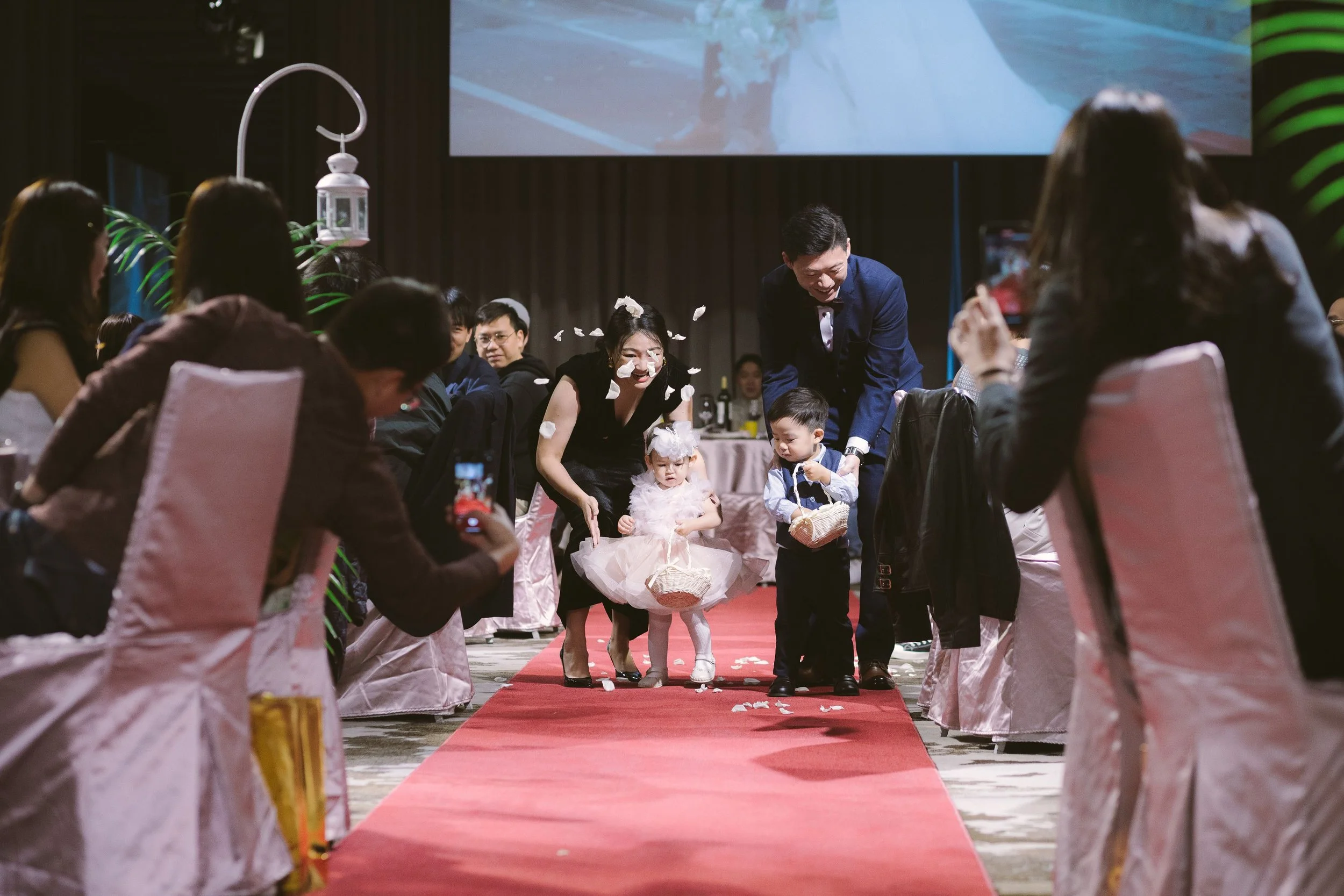 People attending a wedding reception seated on either side of a red carpet, with a newlywed couple and two young children walking down the aisle while flower petals fall around them.