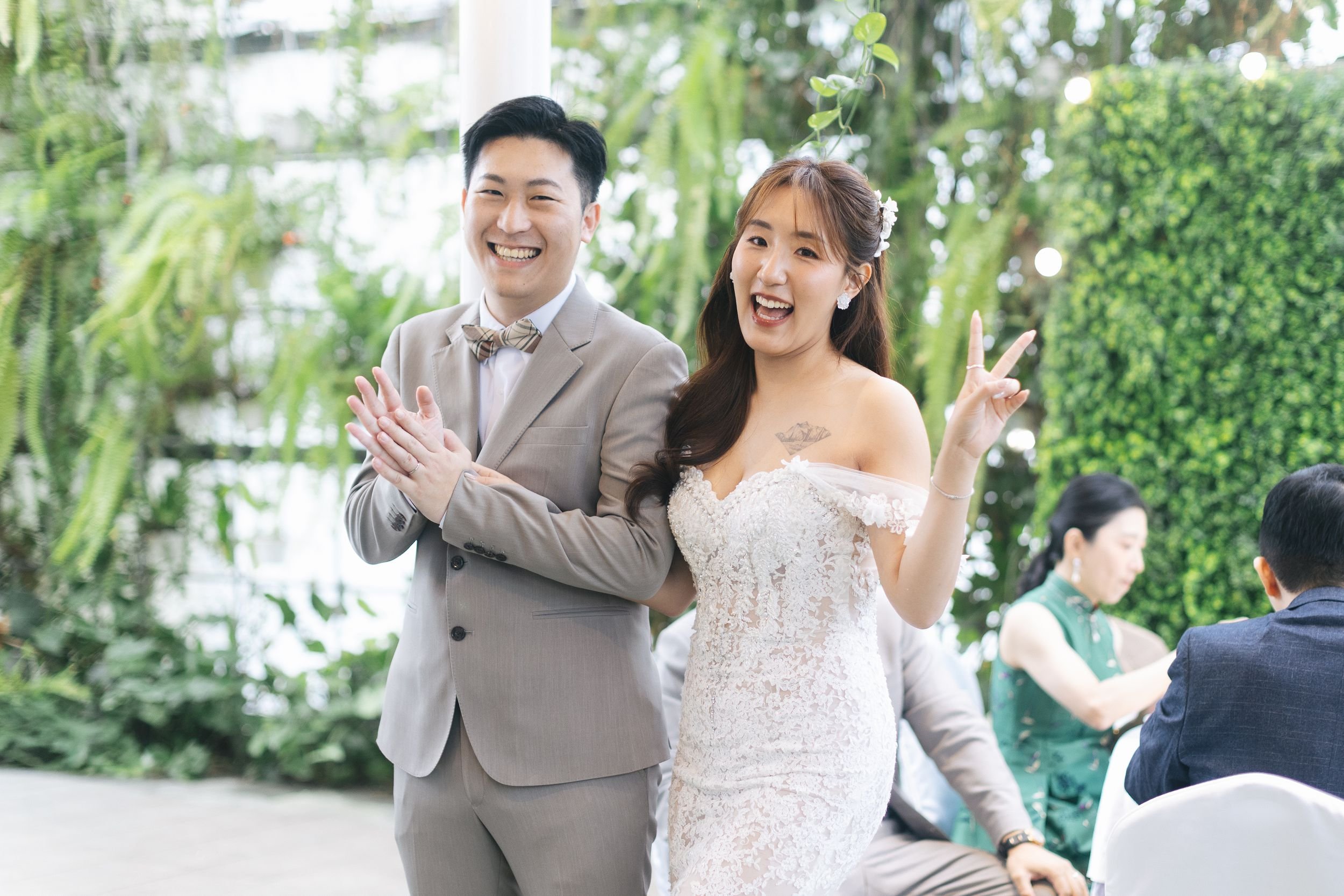 A happy bride and groom at their wedding reception. The bride is making a peace sign with her hand, and the groom is smiling. They are indoors with lush green plants in the background.