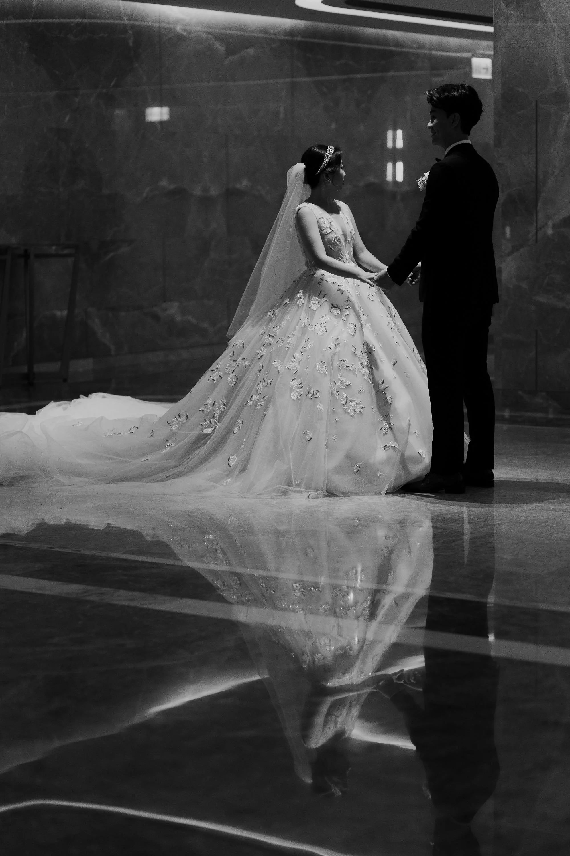 A black and white photo of a bride and groom holding hands during their wedding ceremony. The bride wears a detailed wedding gown with a long train and a veil, while the groom wears a dark suit. They are facing each other in front of a dark, marble-l