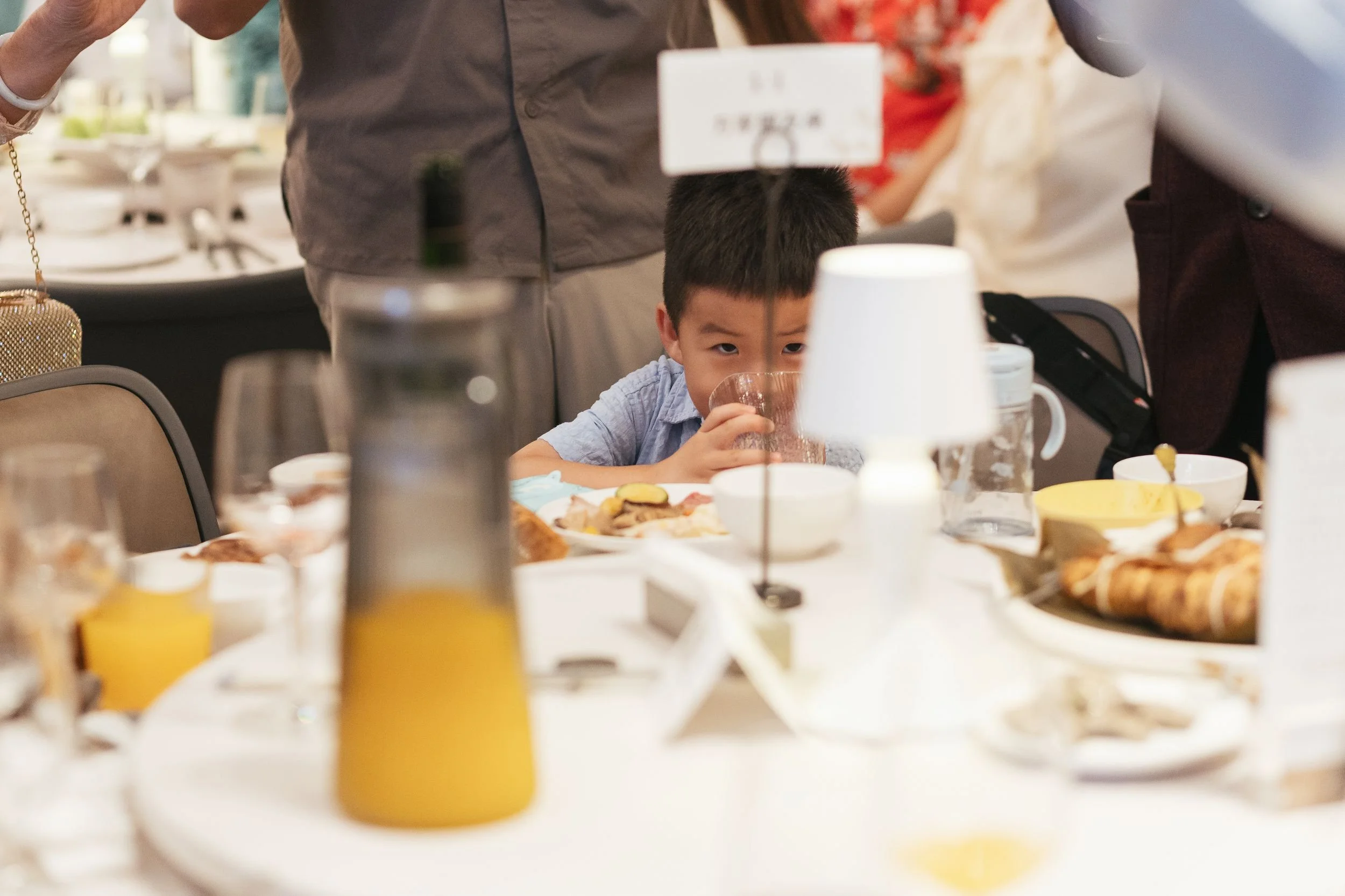 Young boy drinking from a glass at a table during a meal with plates of food, drinks, and condiments, surrounded by adults in a dining setting.