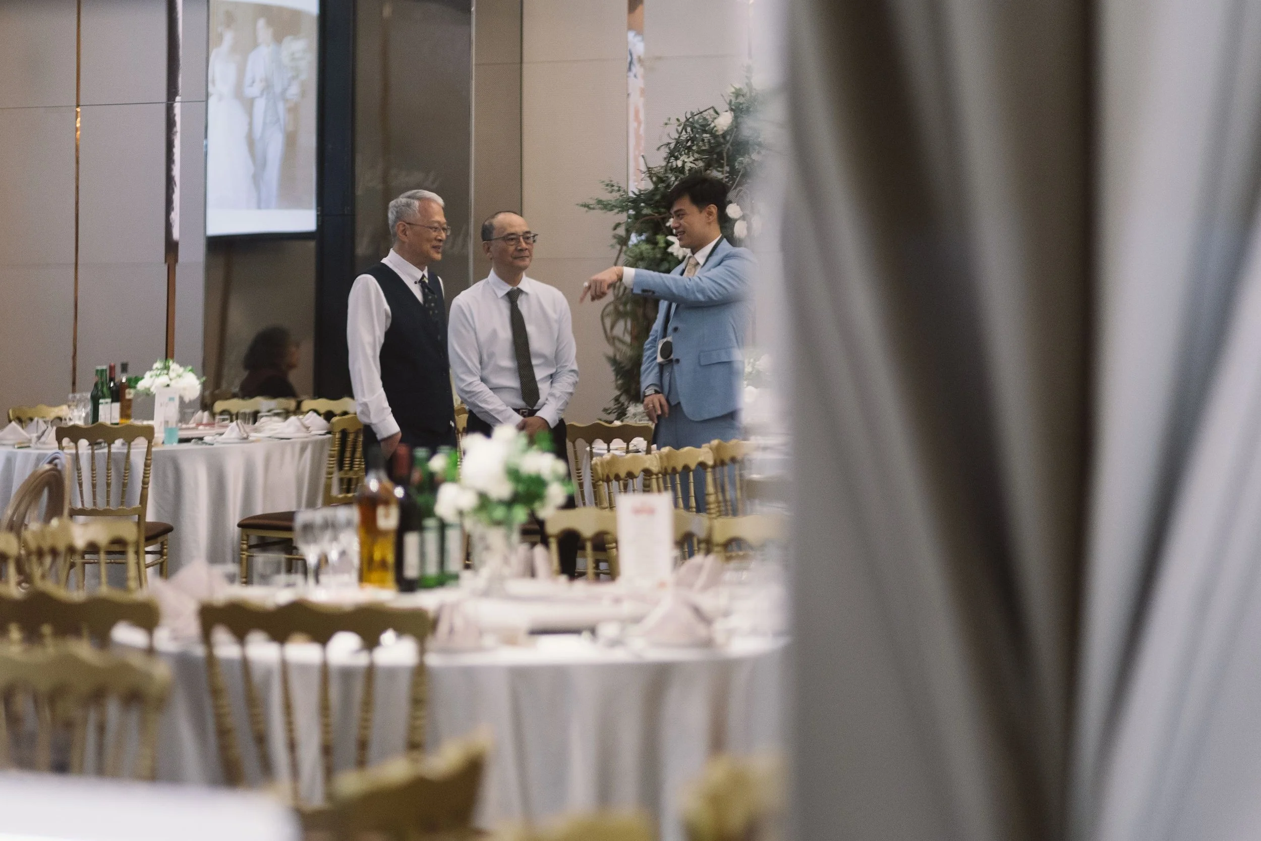 Three men dressed in formal attire are engaged in conversation at a decorated event space with tables set for a meal, floral arrangements, and bottles of beverages, viewed through a partially obscuring foreground.