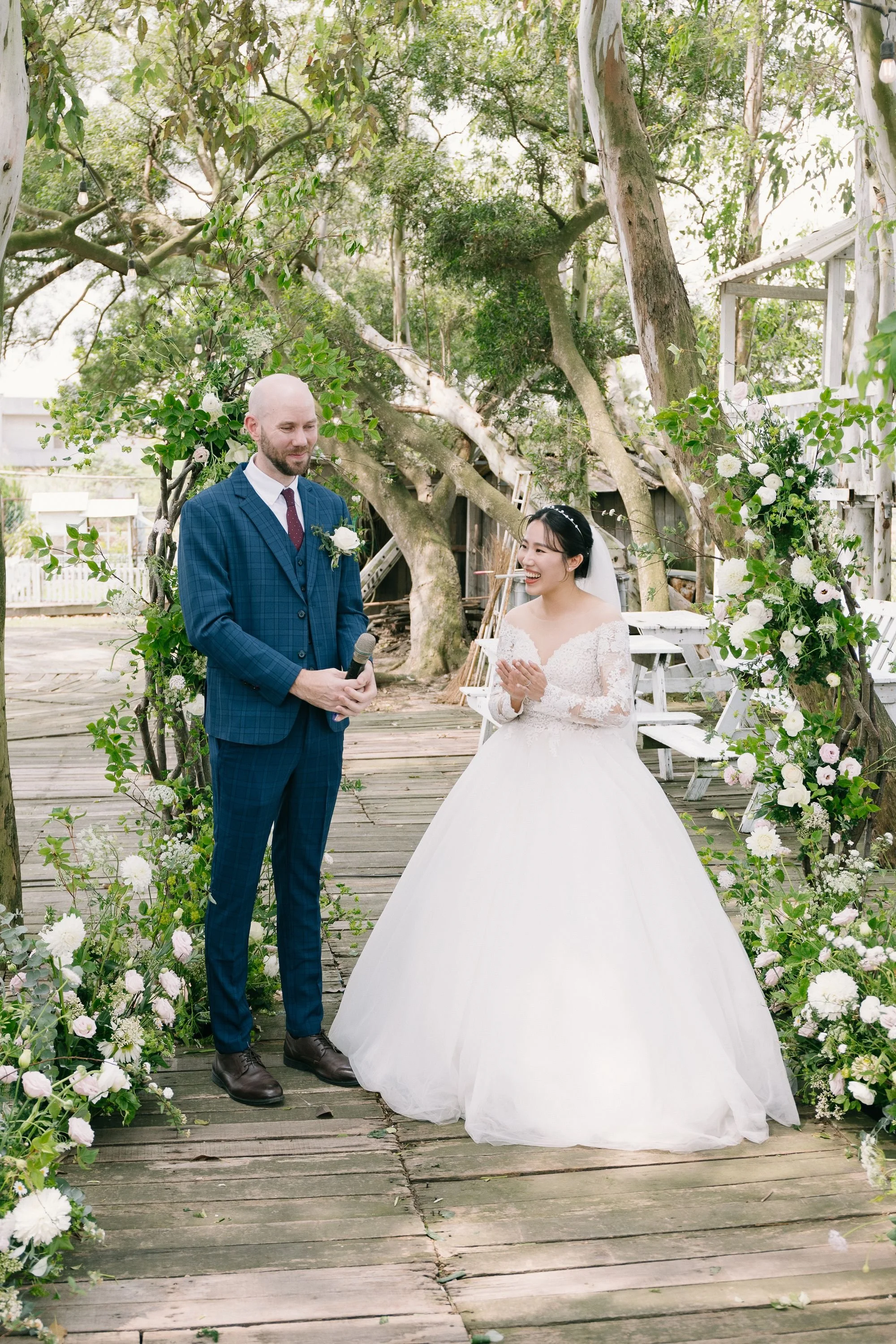 A bride and groom stand under decorated archway outdoors with trees in the background, during a wedding ceremony. The groom wears a blue plaid suit, and the bride wears a white lace wedding dress and looks at him smiling.