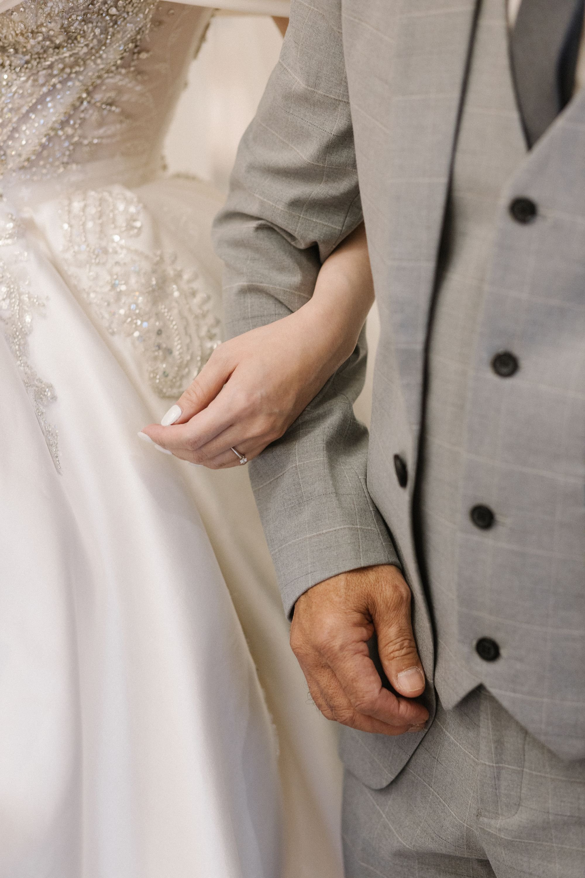 A close-up of a bride and groom holding hands, with the bride wearing a detailed beaded dress and the groom in a light grey suit.