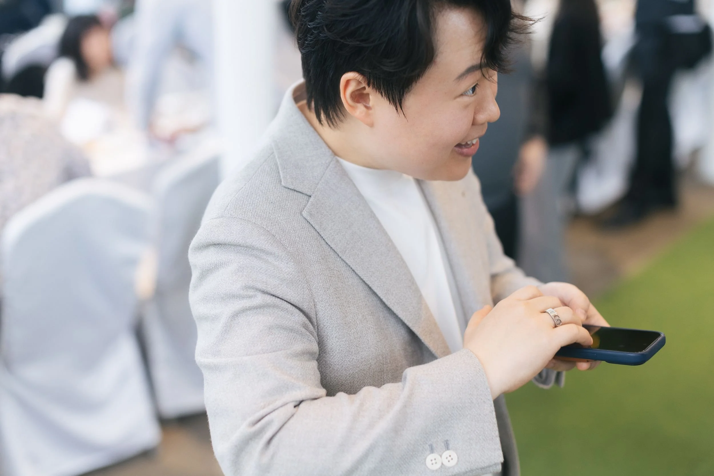 A woman with short dark hair wearing a light-colored blazer is looking at her phone and smiling. She is in a social setting with other people in the background.