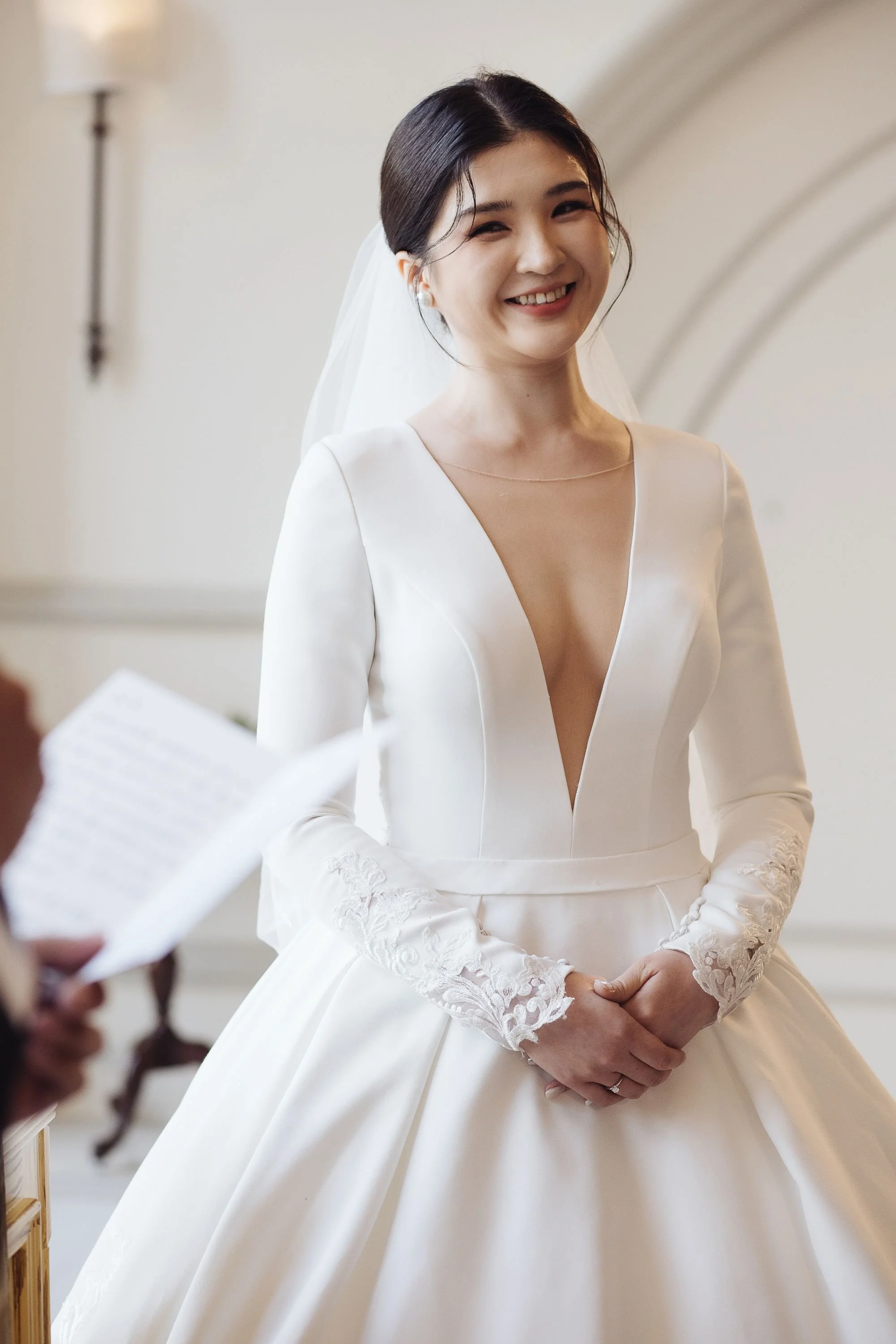 A smiling bride with dark hair, wearing a white wedding dress with lace sleeves and a sheer neckline, standing in an indoor setting.