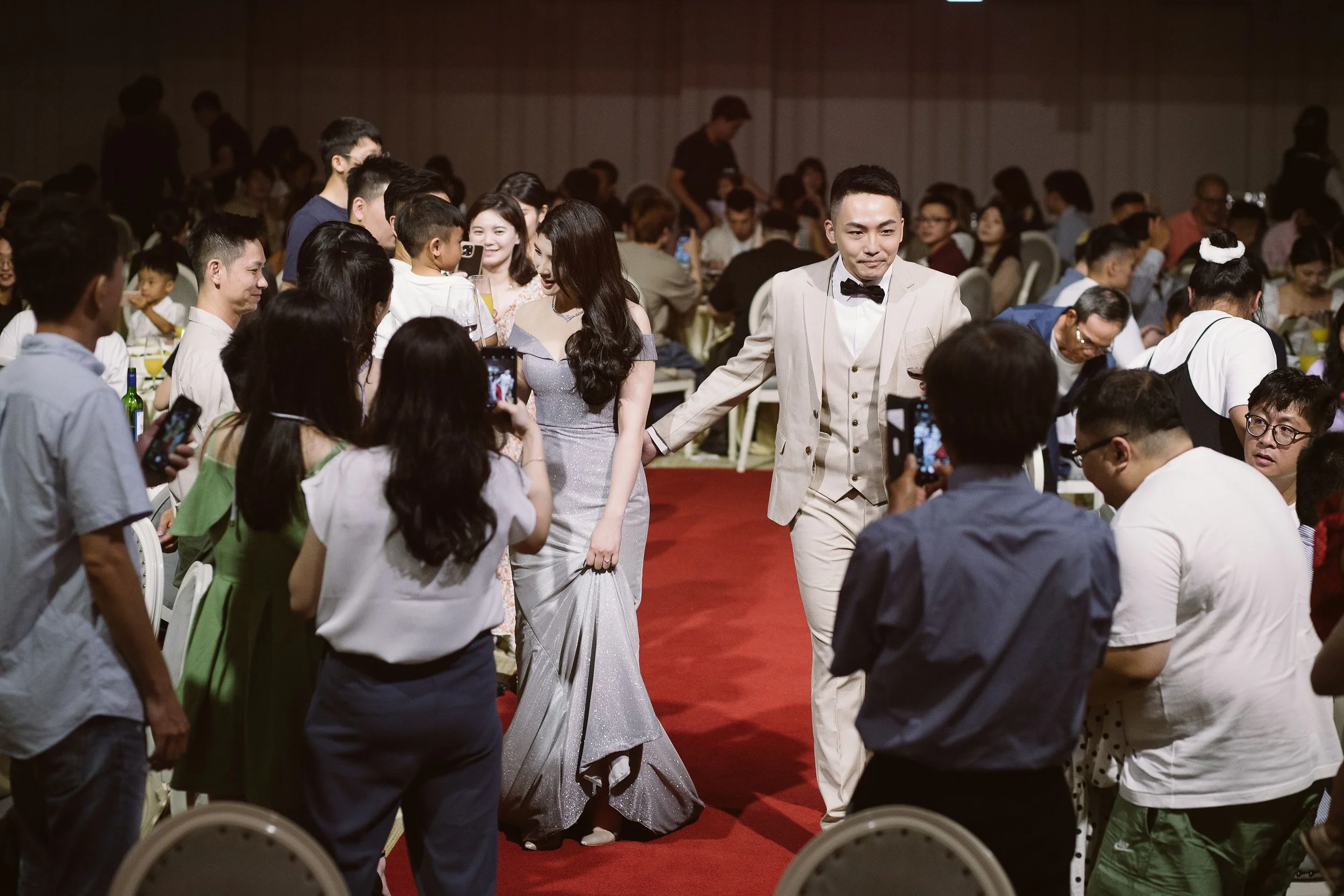 A bride and groom walking down a red carpet surrounded by seated guests at a wedding reception.