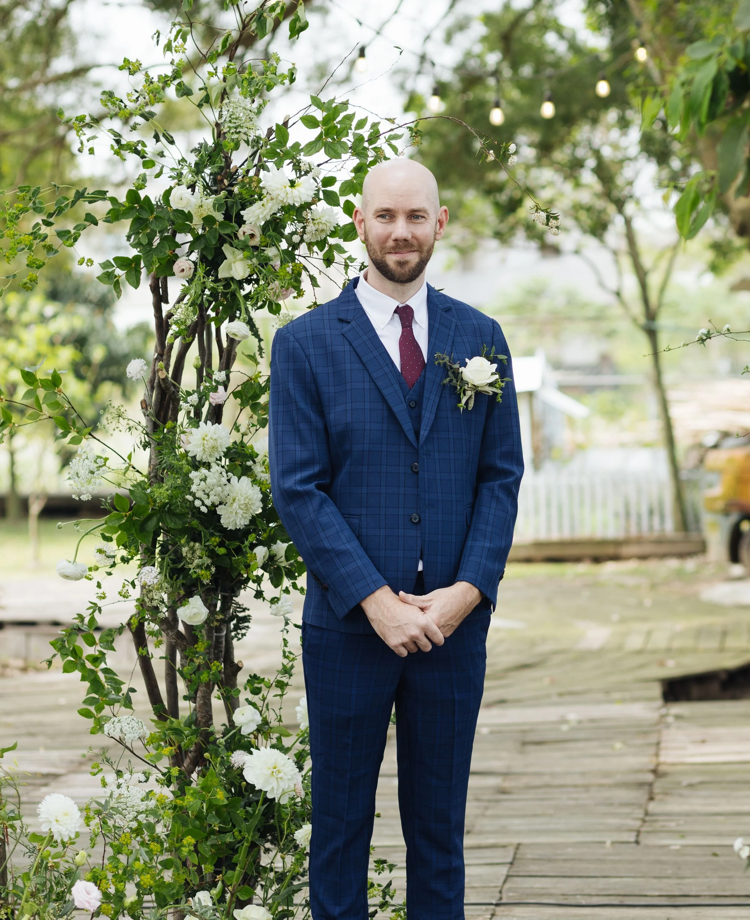 A bald man in a blue plaid suit with a white shirt and maroon tie, standing outdoors on a wooden walkway with greenery and flowers behind him, with tree branches and string lights overhead.