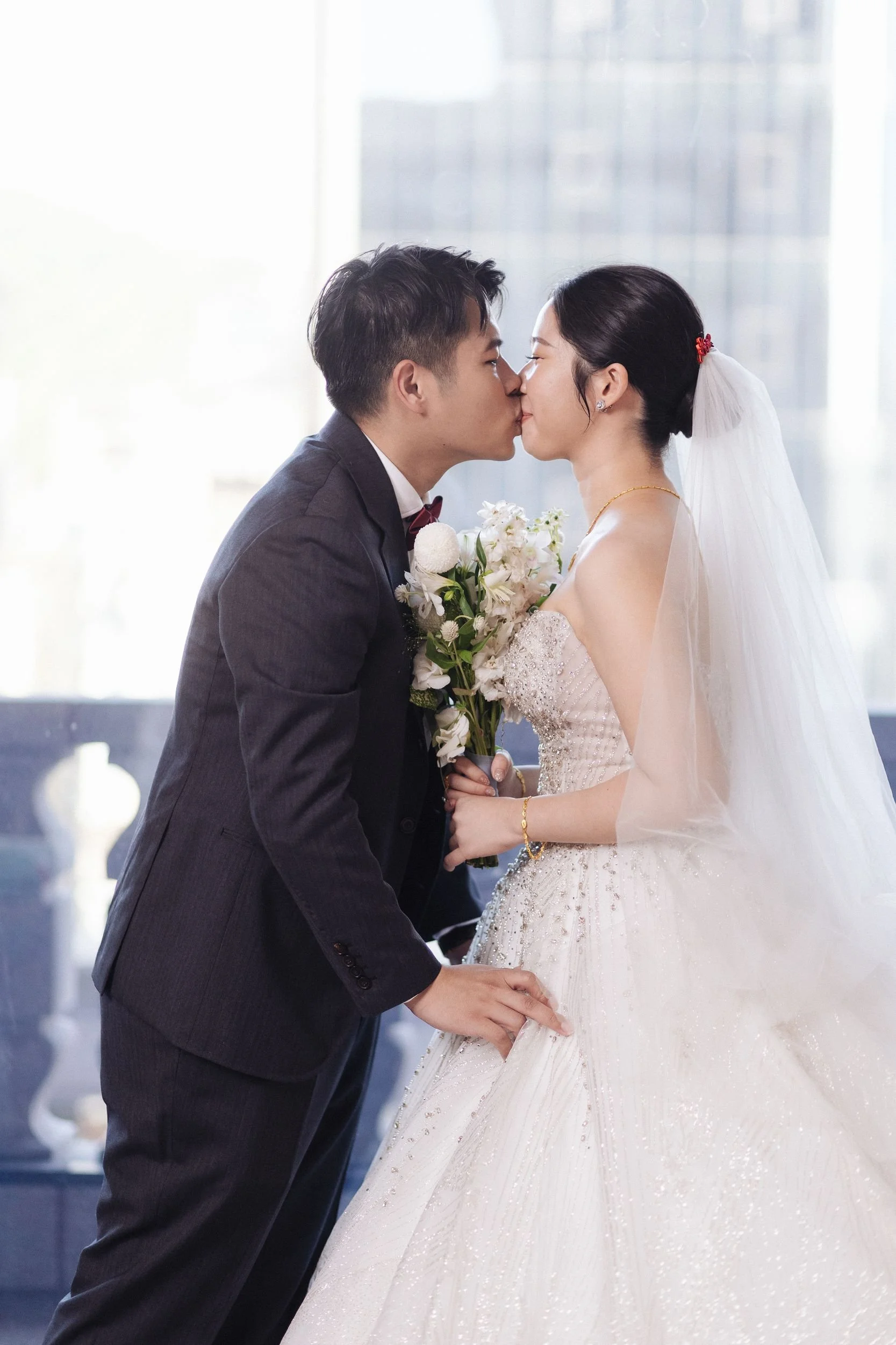 A groom and bride are sharing a kiss at their wedding. The groom is wearing a dark suit with a bow tie, and the bride is in a white wedding gown with a veil. She holds a bouquet of white flowers. They are standing close to each other in a brightly li