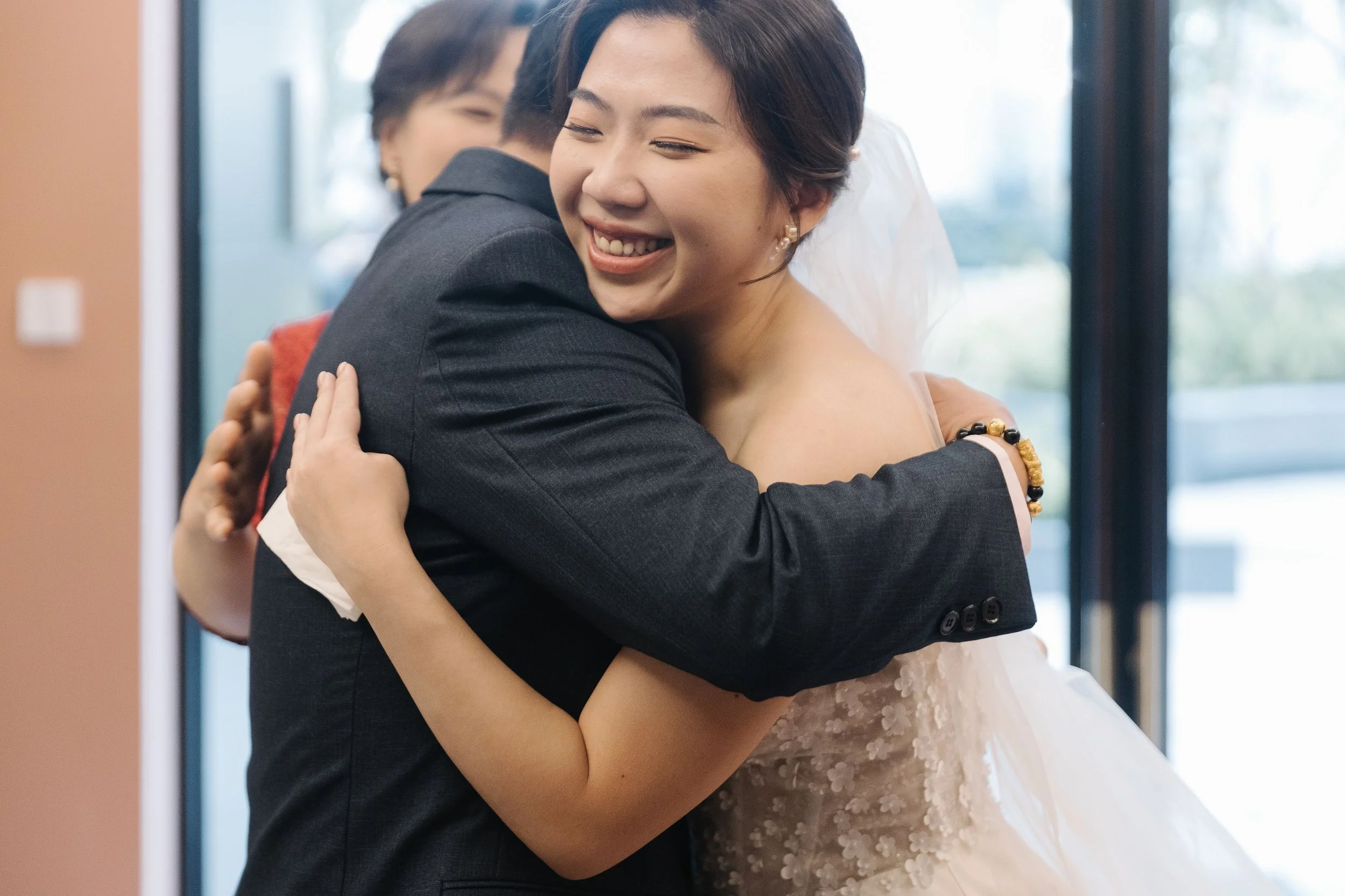 A woman in a wedding dress and a man in a dark suit hugging happily in an indoor setting with large windows.