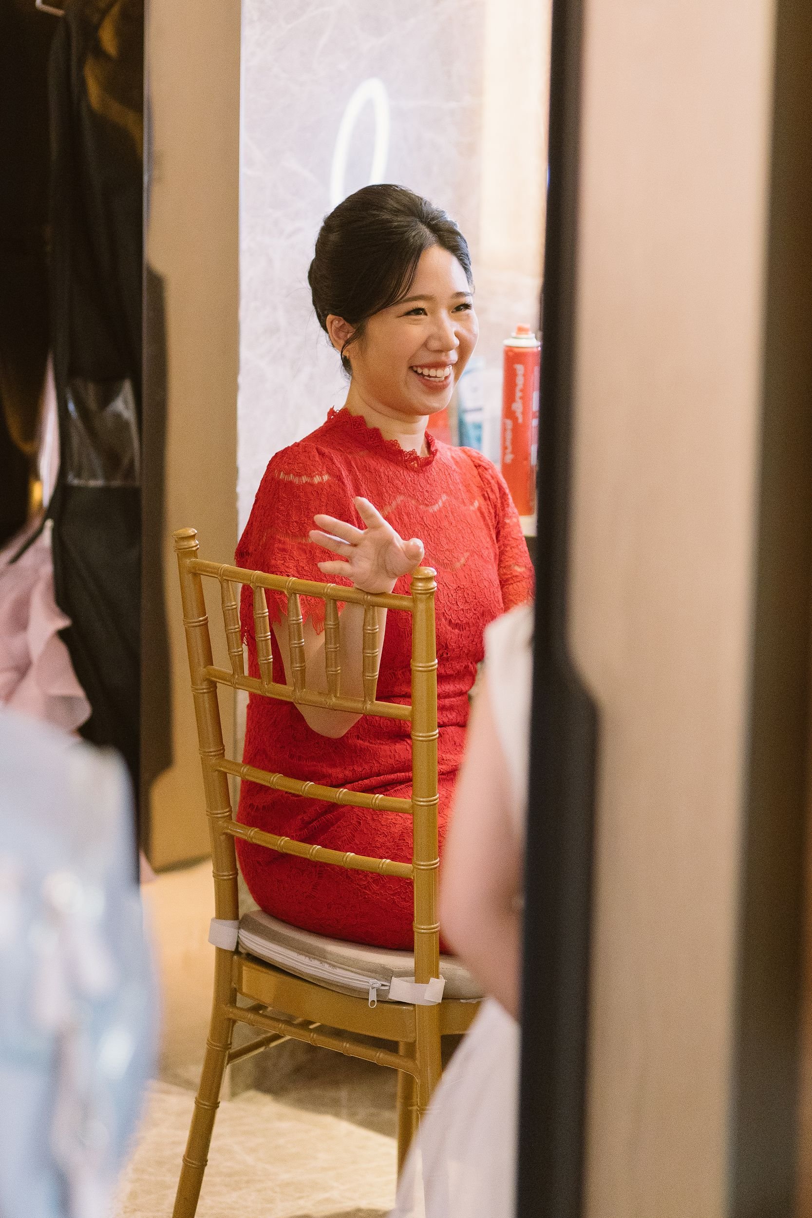 A woman in a red lace dress smiling and waving while sitting on a gold-colored chair, reflected in a mirror.