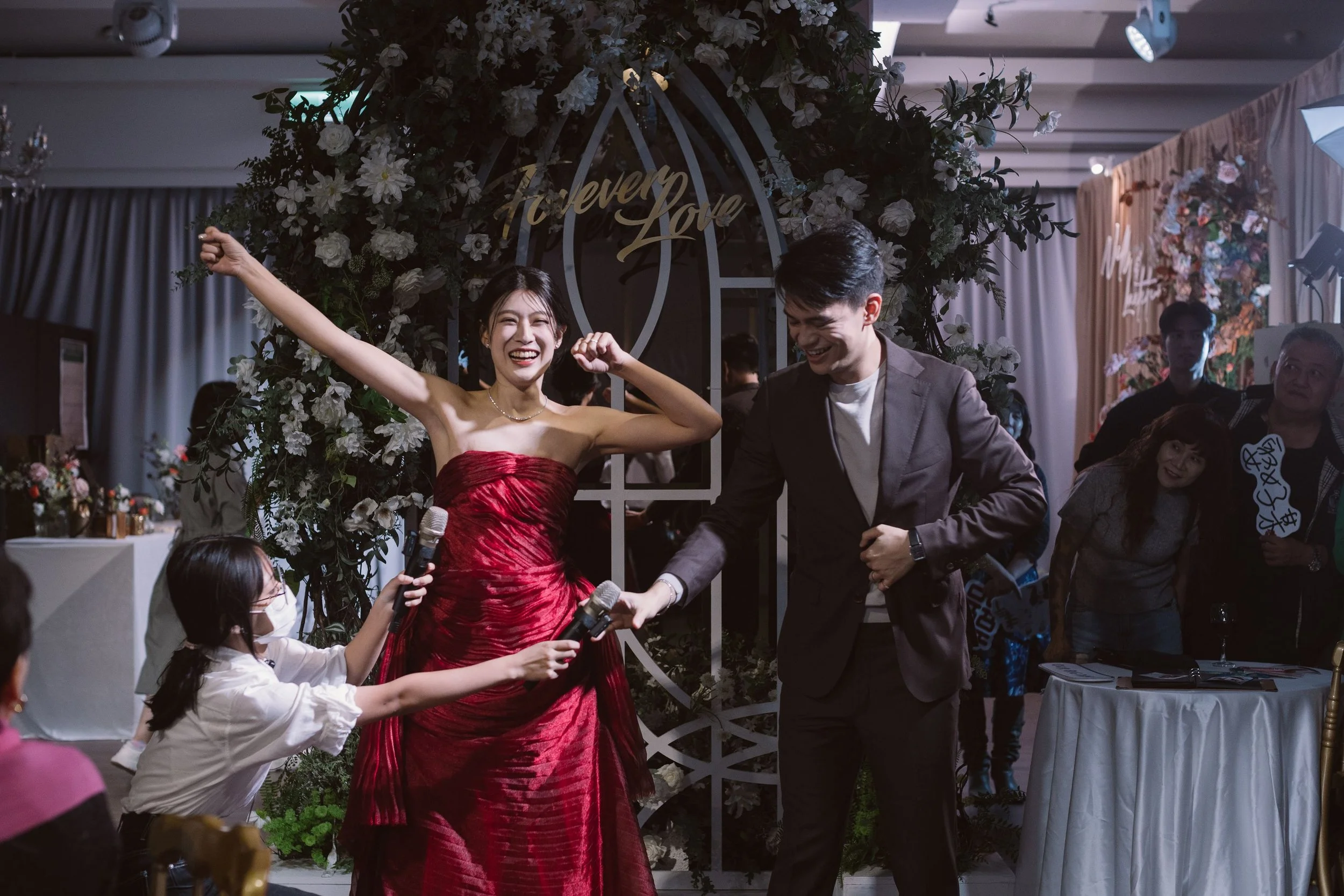 Joyful woman in elegant red dress celebrating with others at a wedding or party, holding microphones, under a floral arch with a sign that says 'Every Love'.