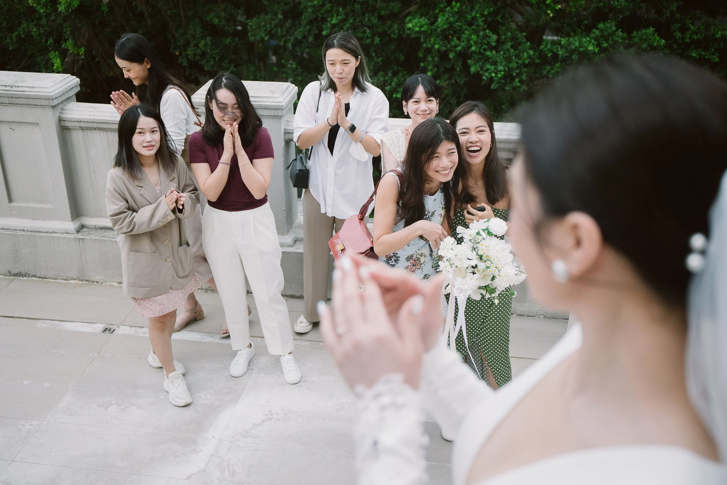 Eight women standing outdoors near a stone railing, smiling, and clapping their hands. Two women are holding flower bouquets, and others appear to be praying or celebrating. The background is filled with green trees.