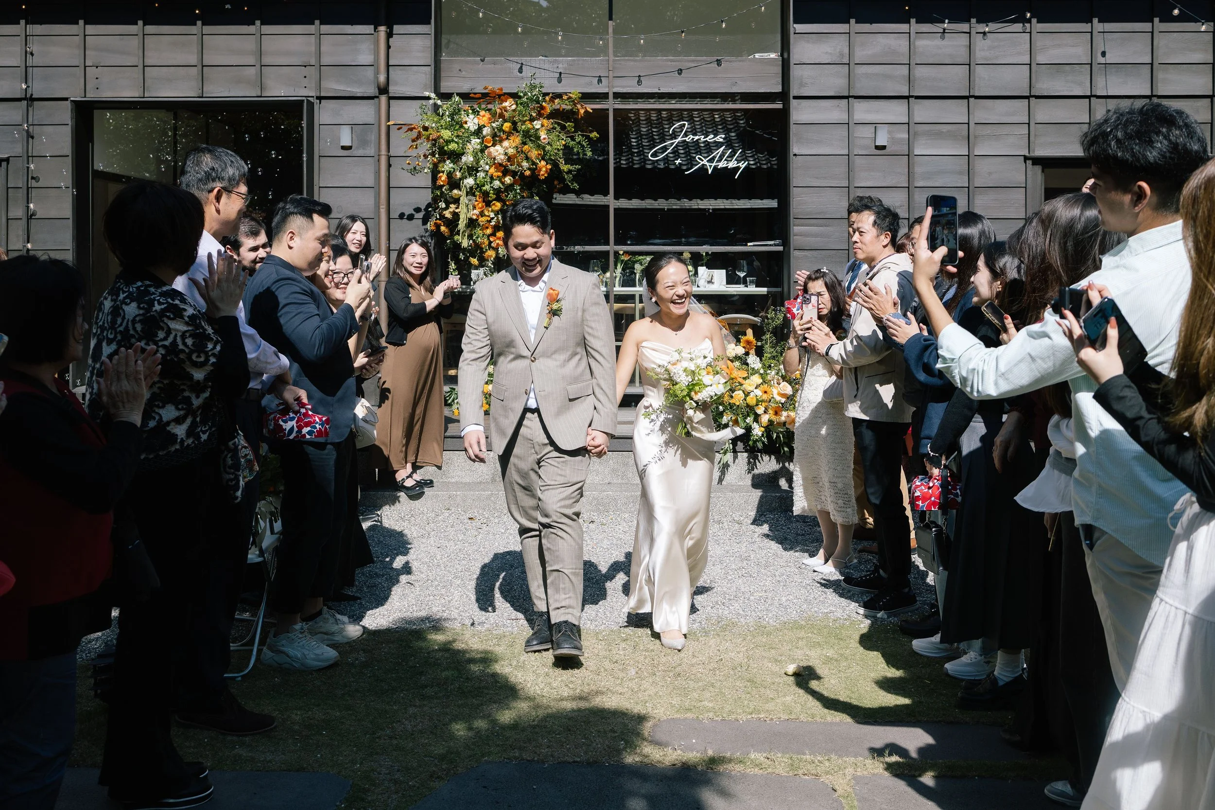 A wedding scene with a bride and groom walking through a aisle lined with guests, outside a building with a sign that says 'Jones Abby.' The bride is holding a bouquet, and the groom is wearing a beige suit. Guests are clapping, smiling, and taking p