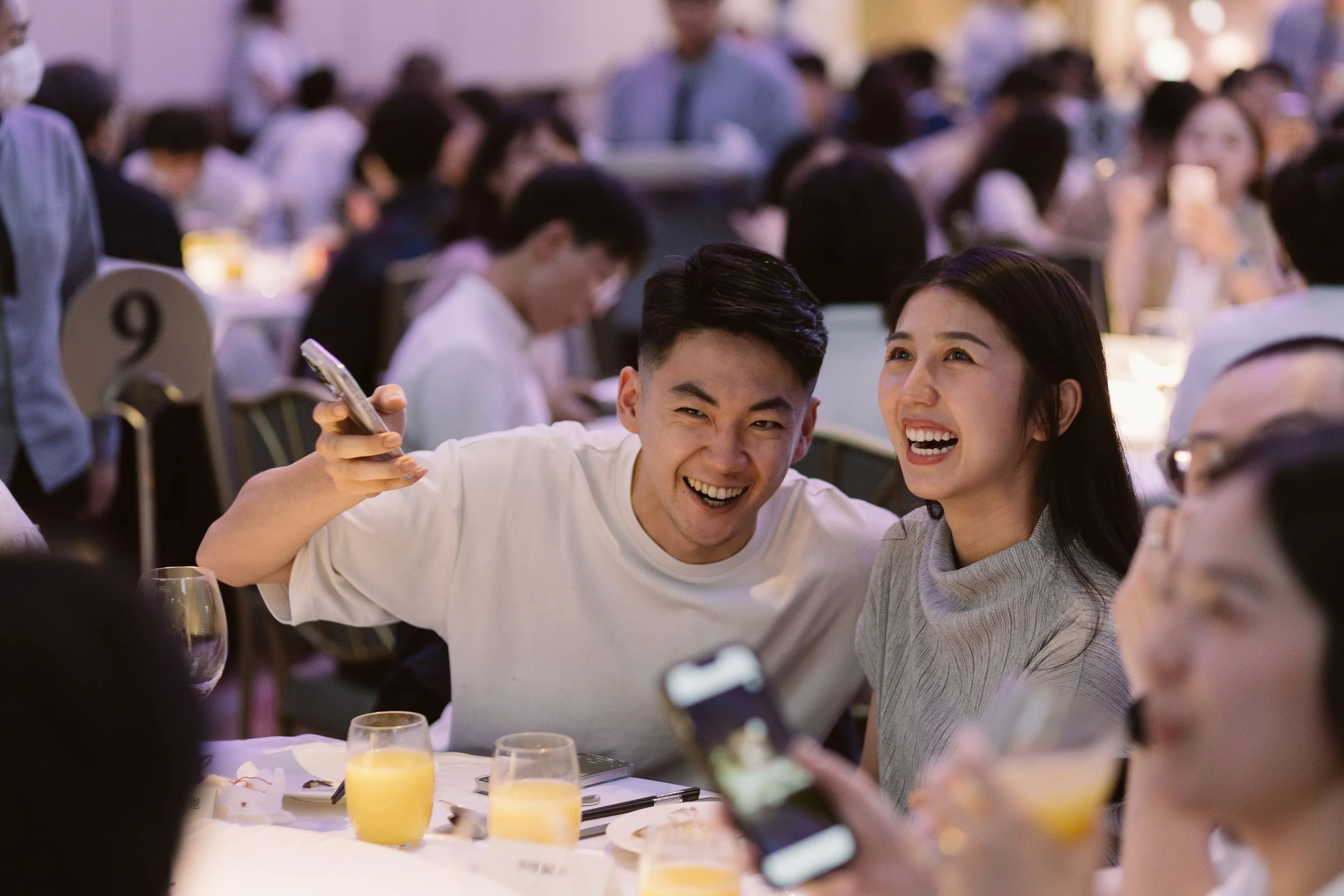 A group of people at a banquet or celebration, sitting around a dining table with glasses of orange juice, smiling and enjoying each other's company in a brightly lit event space.