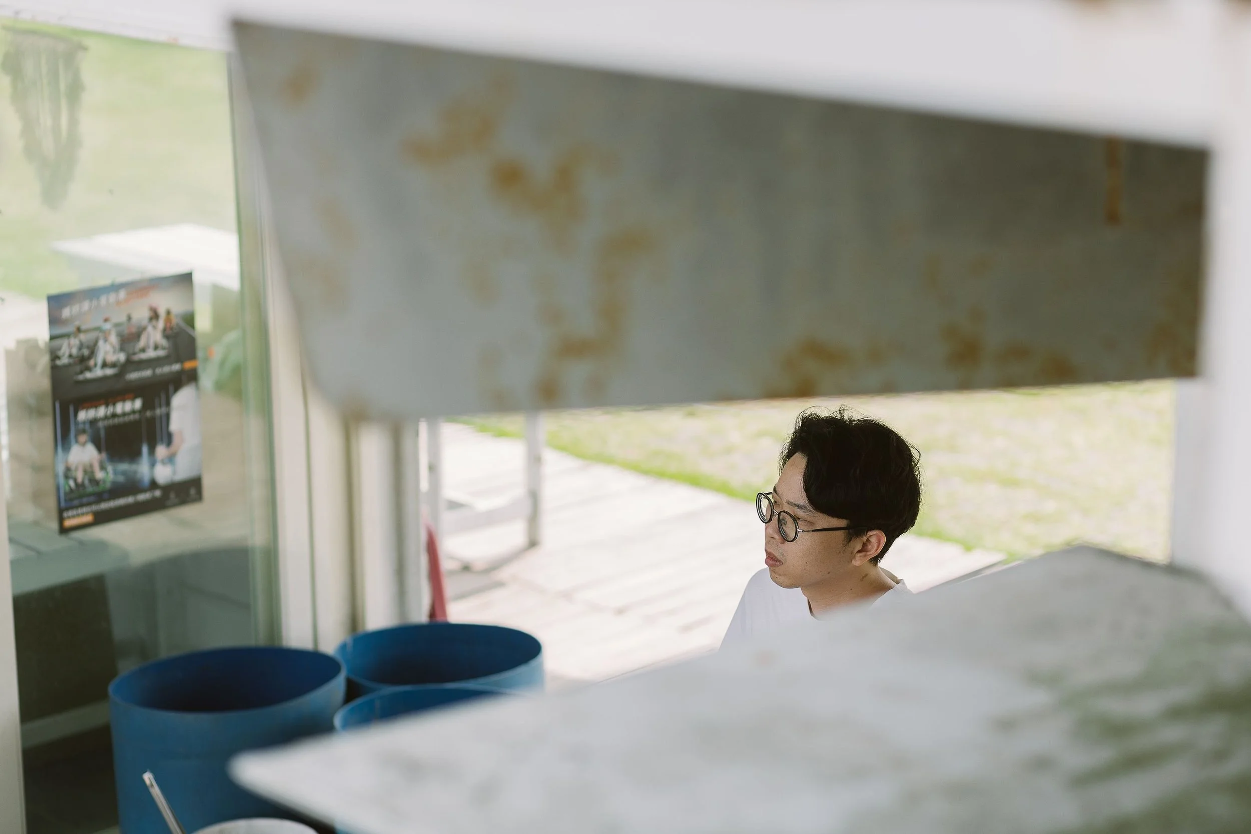 A person with glasses looking downward, seen through a partially opened window or partition in an outdoor setting.