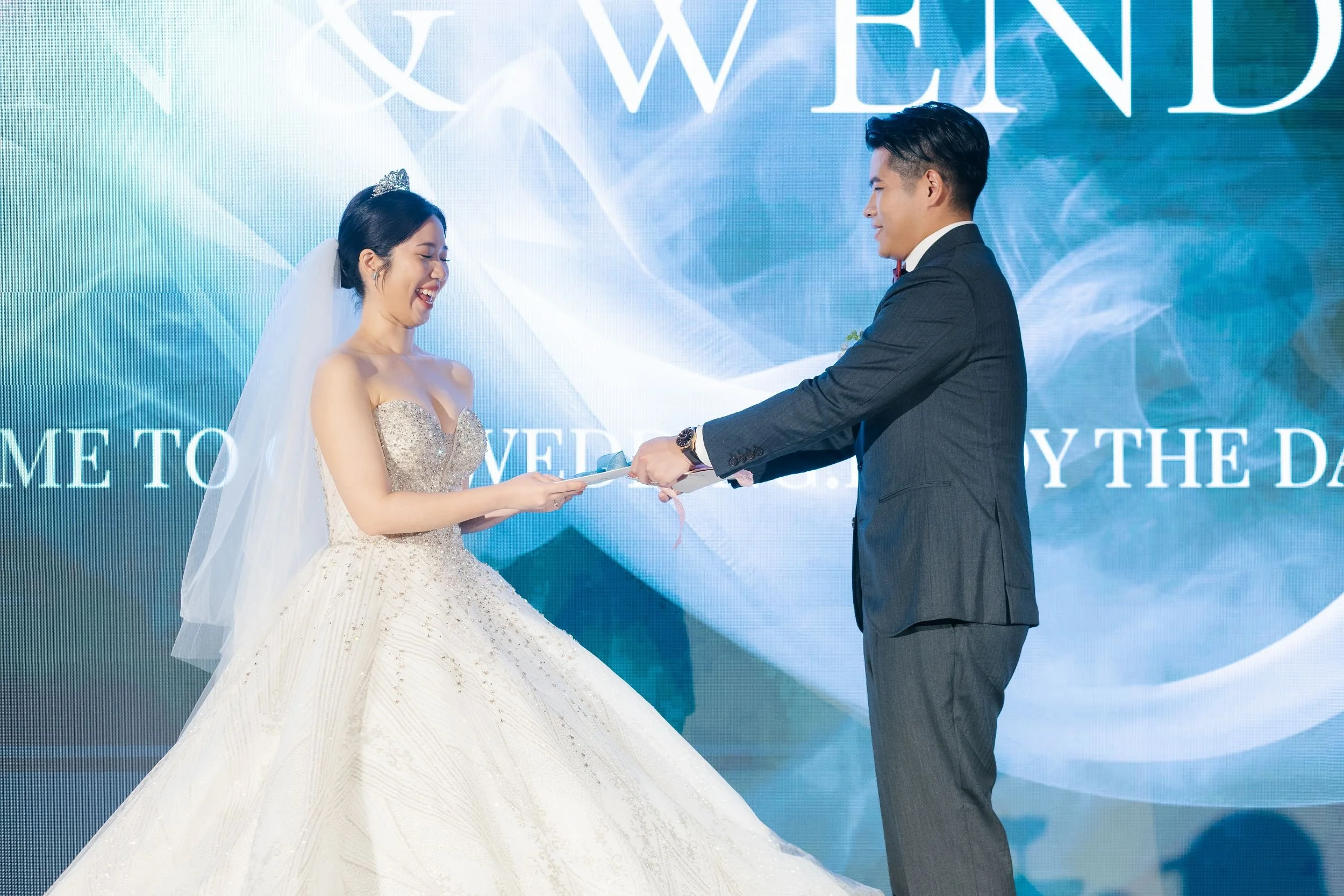 A bride and groom are standing in front of a blue screen with text, during their wedding ceremony. The bride is wearing a strapless white wedding gown with intricate beading and has a veil and a tiara. The groom is in a dark suit with a white shirt a