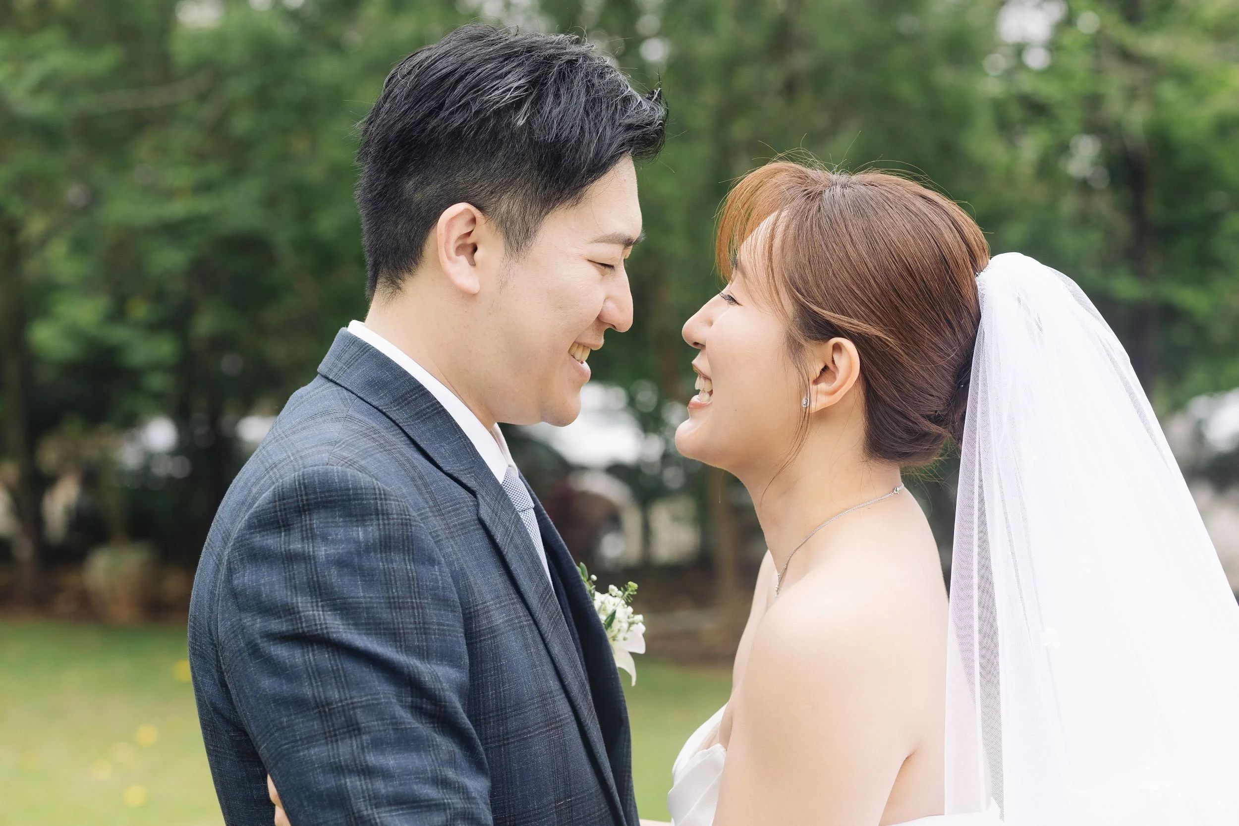 A newlywed couple is smiling and touching foreheads outdoors, with a green, leafy background.
