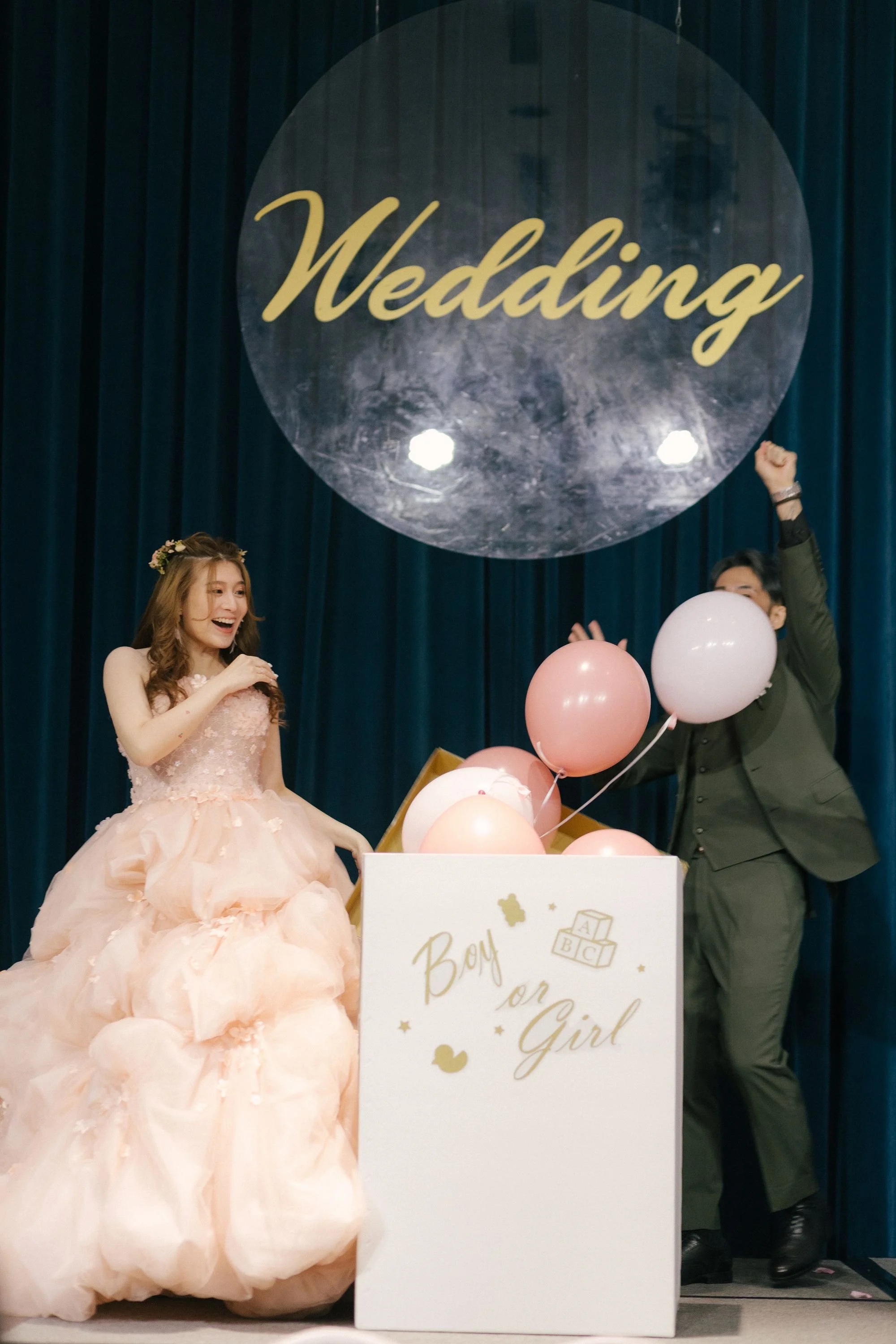 A woman in a pink gown sits on a stage next to a man in a suit. They are celebrating a gender reveal, with balloons and a sign that says 'Boy or Girl' and 'Baby on the way.'