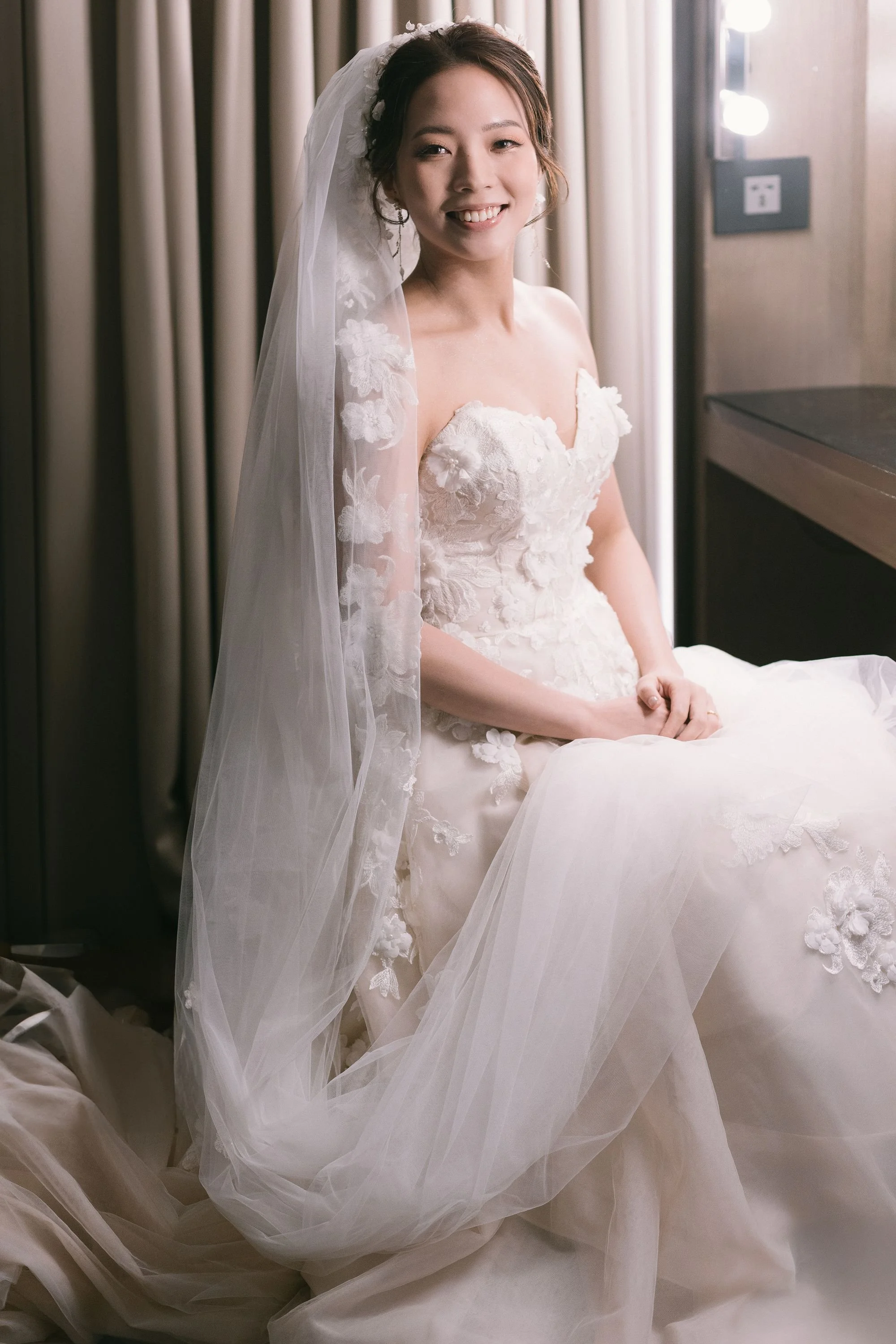 A young woman in a wedding dress with lace floral details and a veil, smiling and sitting on the floor in a dressing room with beige curtains and a few lights.
