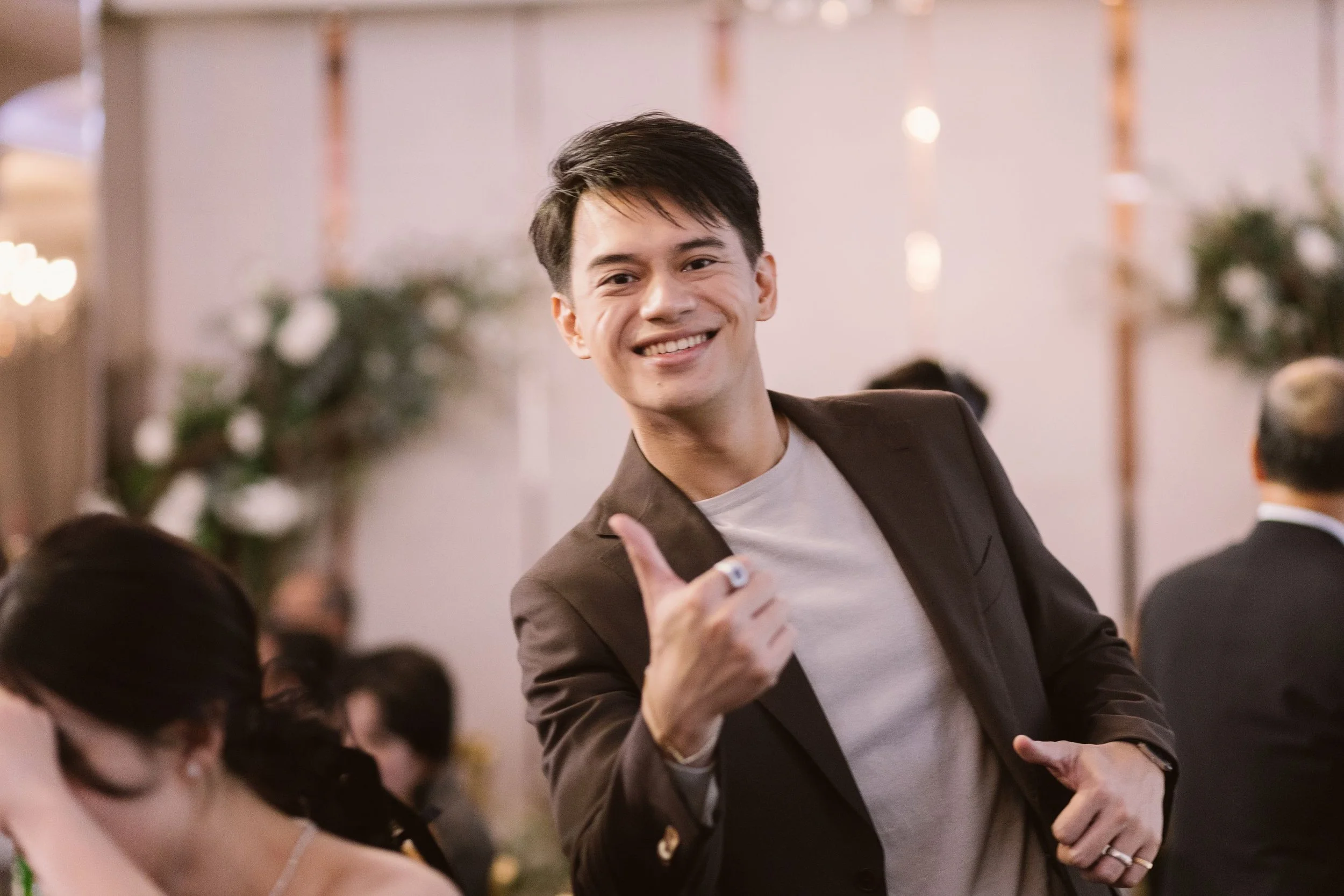 A young man in a gray shirt and brown blazer smiling and giving a thumbs-up at a social gathering indoors, with blurred guests and decor in the background.