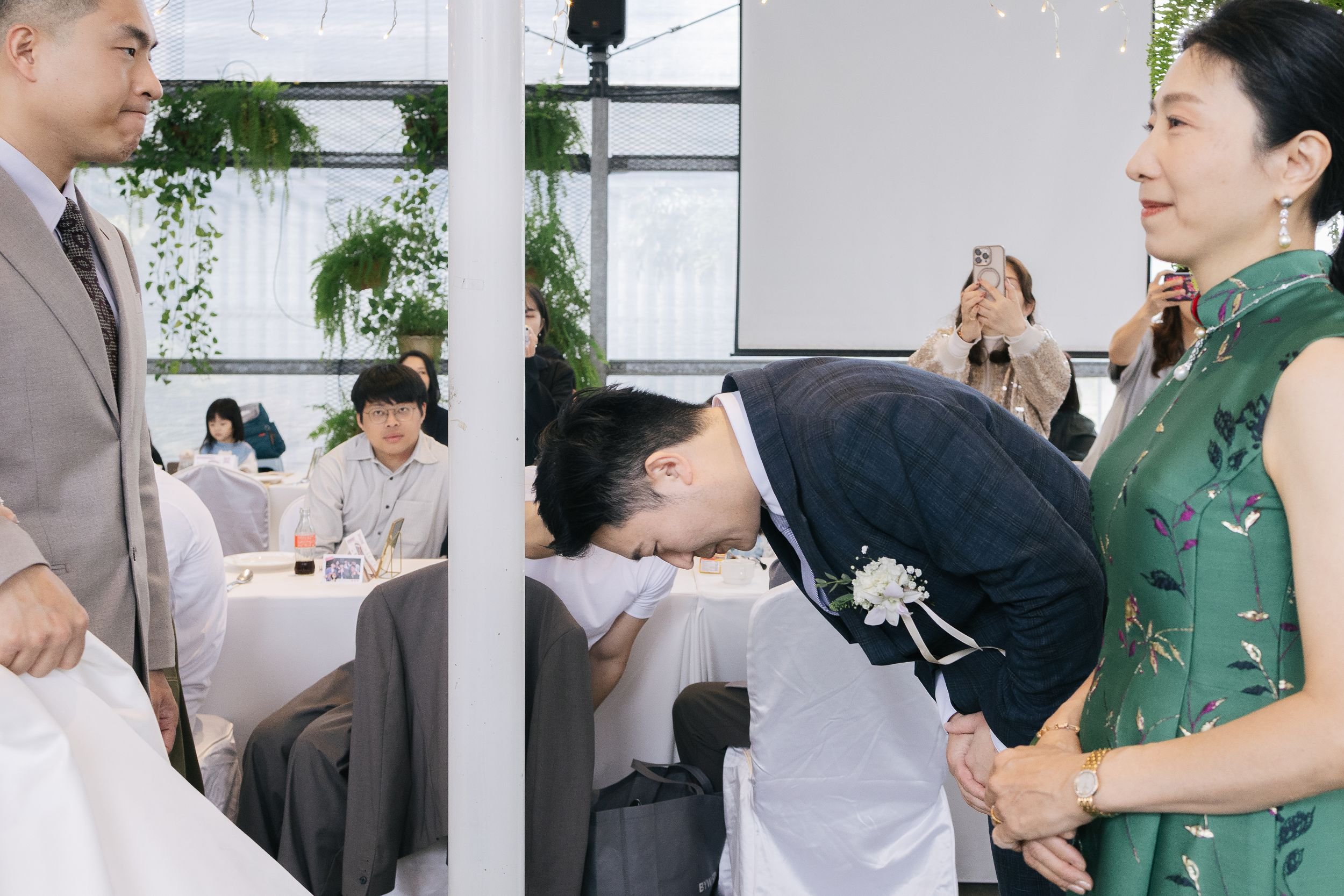 A man in a navy checkered suit and a woman in a green floral dress engaging in a traditional wedding ceremony where the man is bowing his head, and the woman is standing beside him, watching, with other people seated at tables in the background.