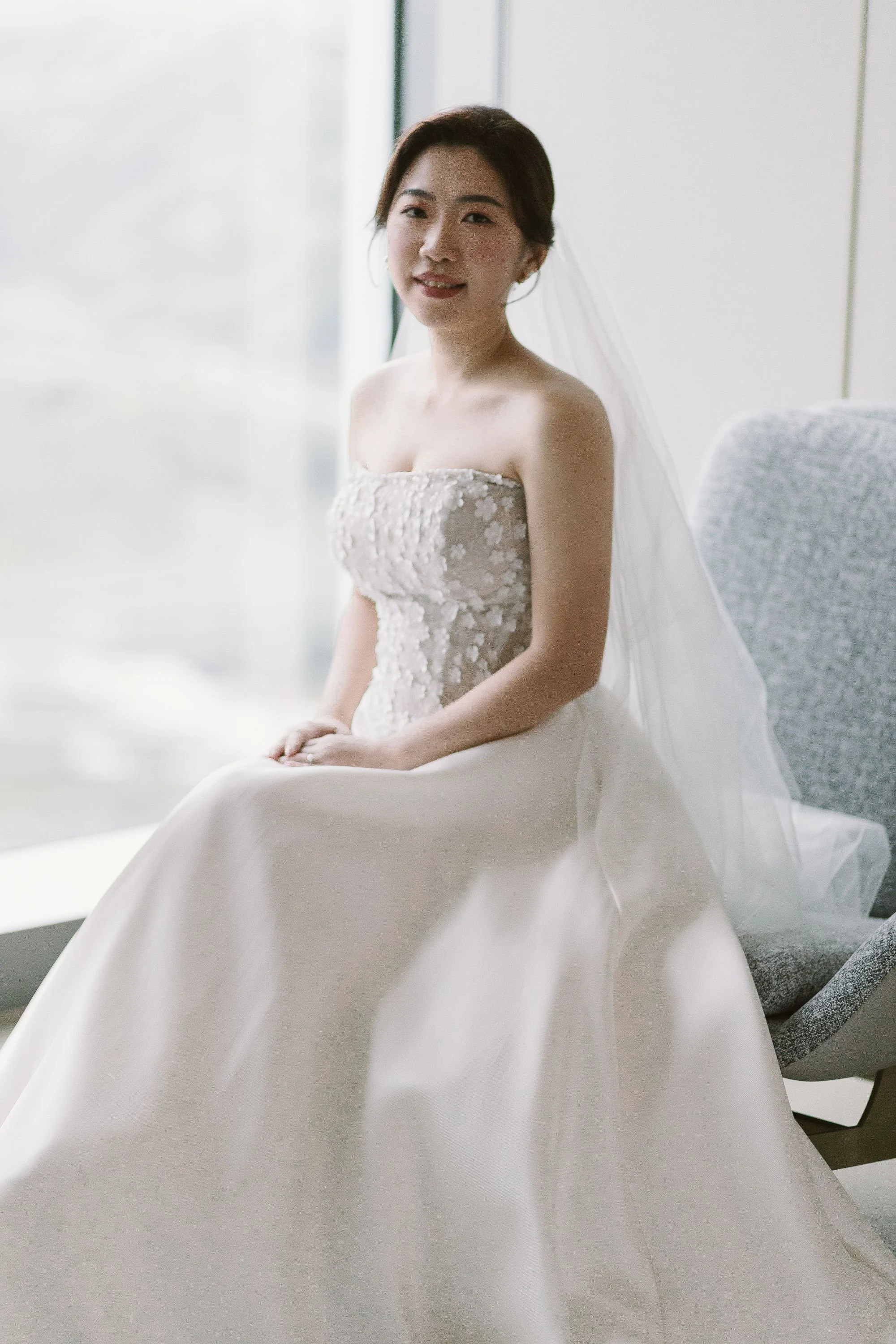 A woman in a wedding dress sitting on a chair, smiling at the camera, with a veil on her head, indoors near a large window.