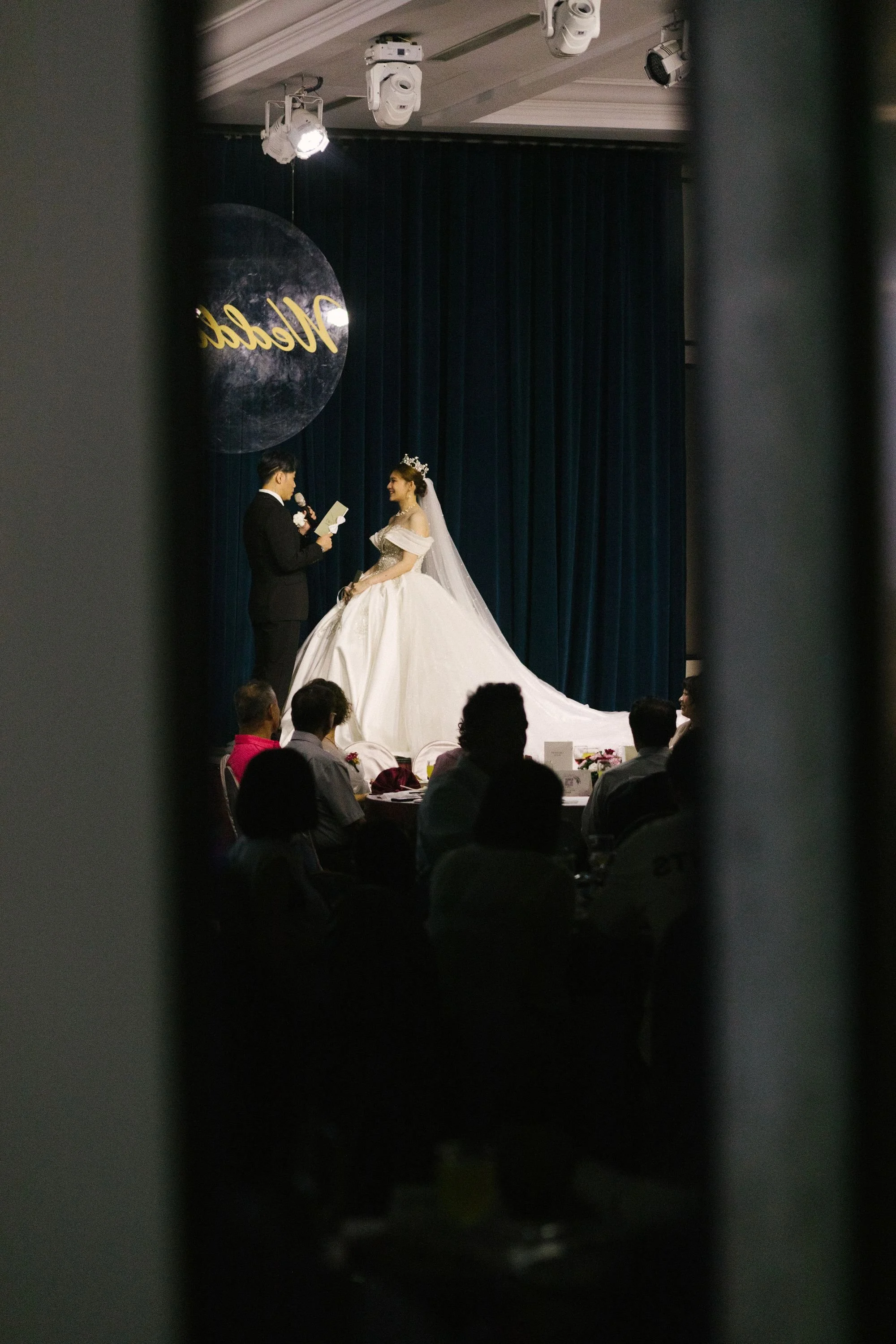 A wedding ceremony in progress, with a bride and groom on stage. The groom is reading from a paper, and the bride is smiling at him, wearing a white wedding dress and tiara. The background features dark curtains and a sign that reads "Wedding".