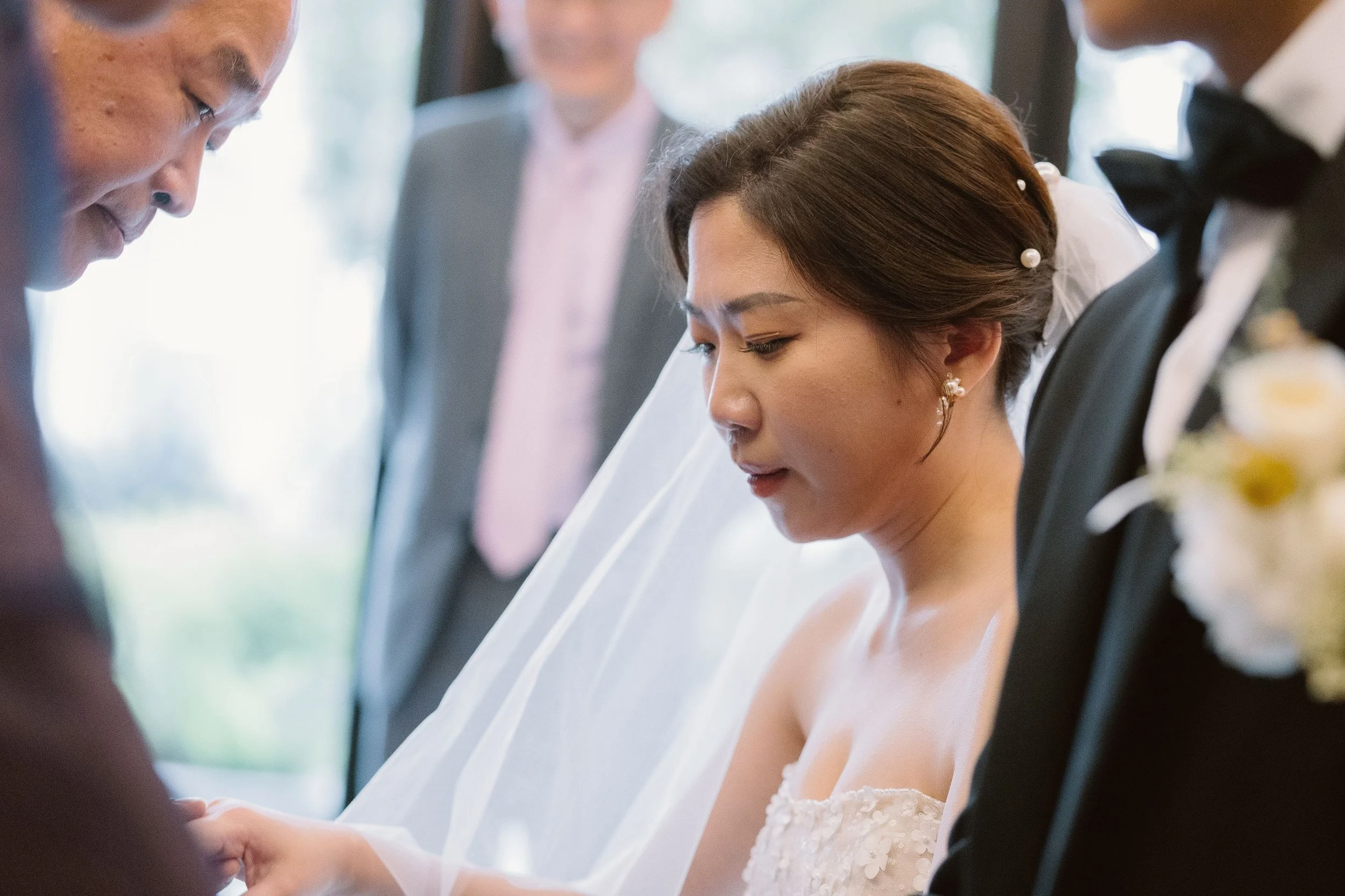 A bride in a white wedding dress and veil is holding hands with her partner during a wedding ceremony, with officiants or witnesses nearby.