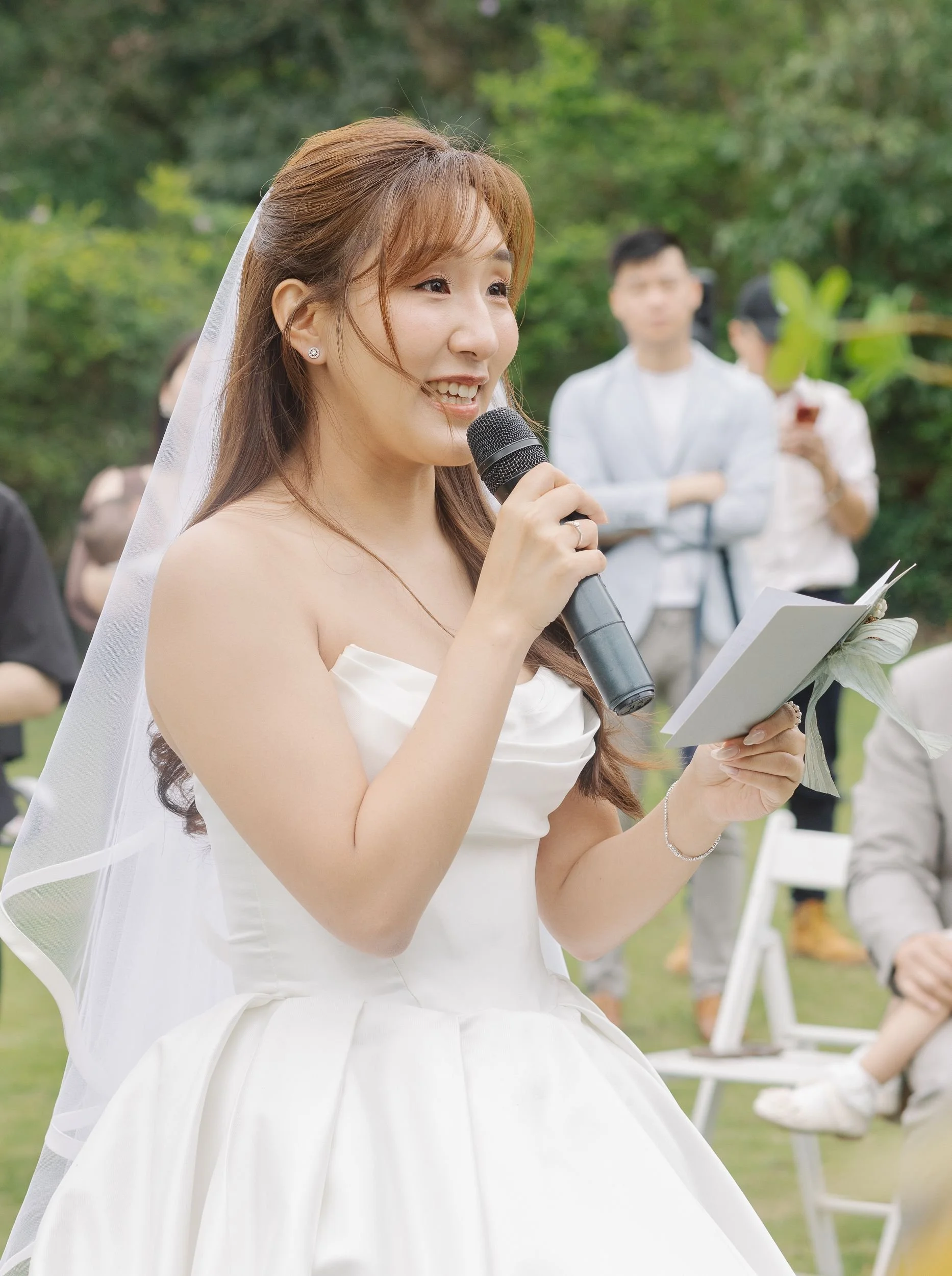 A smiling bride with auburn hair, wearing a white wedding dress and veil, holding a microphone and a small notepad outdoors with blurred guests and green trees in the background.